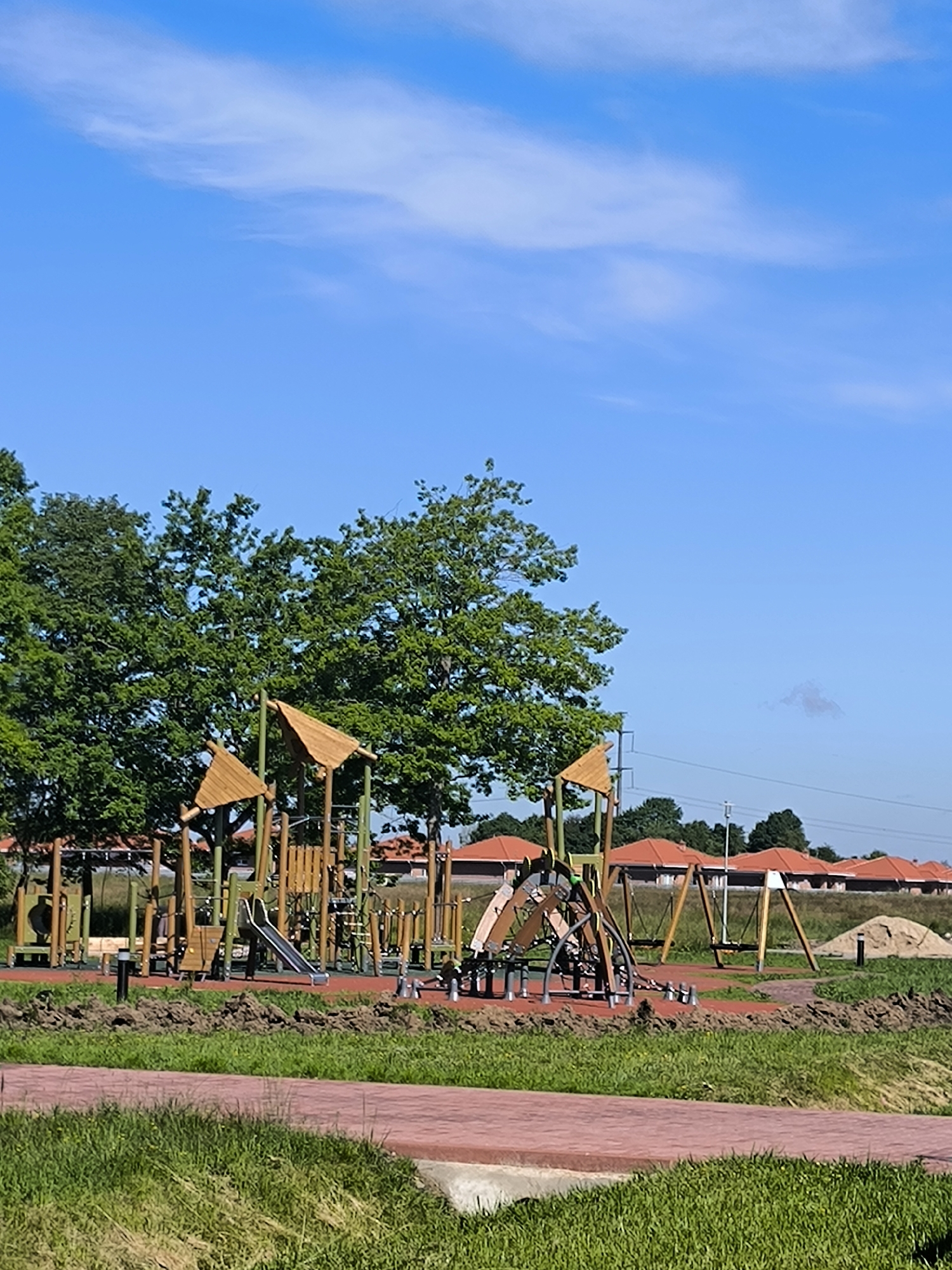 This is a daytime, eye-level, vertical, full shot of a new, modern playground in Laskino, Russia, situated between a green grassy field and a row of houses with terracotta roofs. The playground features a variety of wooden and metal structures, including a slide, swings, and climbing equipment, all set on a red rubberized surface. The ground around the playground is a mix of dirt and grass, suggesting recent construction or landscaping. In the background, mature trees with lush green foliage line the edge of the playground, and beyond them, a clear blue sky with a few wispy clouds stretches overhead. The houses in the distance are uniform in design, with dark tiled roofs and light-colored walls. The scene appears peaceful and under development, with no people visible.