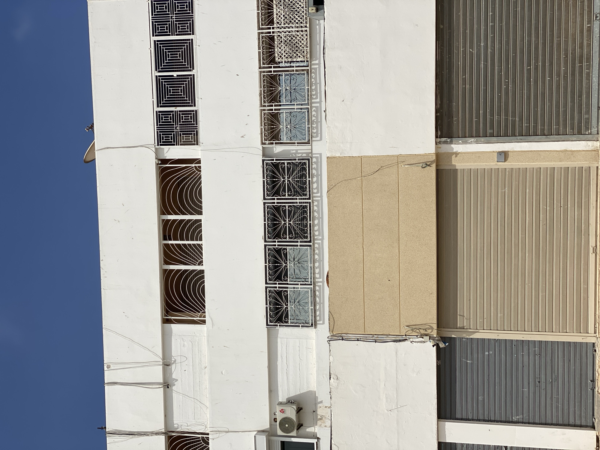 The image shows the facade of a white building in Agadir, Morocco, under a clear blue sky. The building has multiple windows with decorative metal grilles. Some windows have curved, fan-like patterns, while others feature geometric square designs. Below the windows, the building transitions to a beige section with horizontal paneling, and further down, it features large, closed metal roller doors in grey and beige. An air conditioning unit is visible on the left side of the building. Wires and conduits are attached to the exterior of the building. The overall scene depicts a commercial or residential structure in an urban setting.
