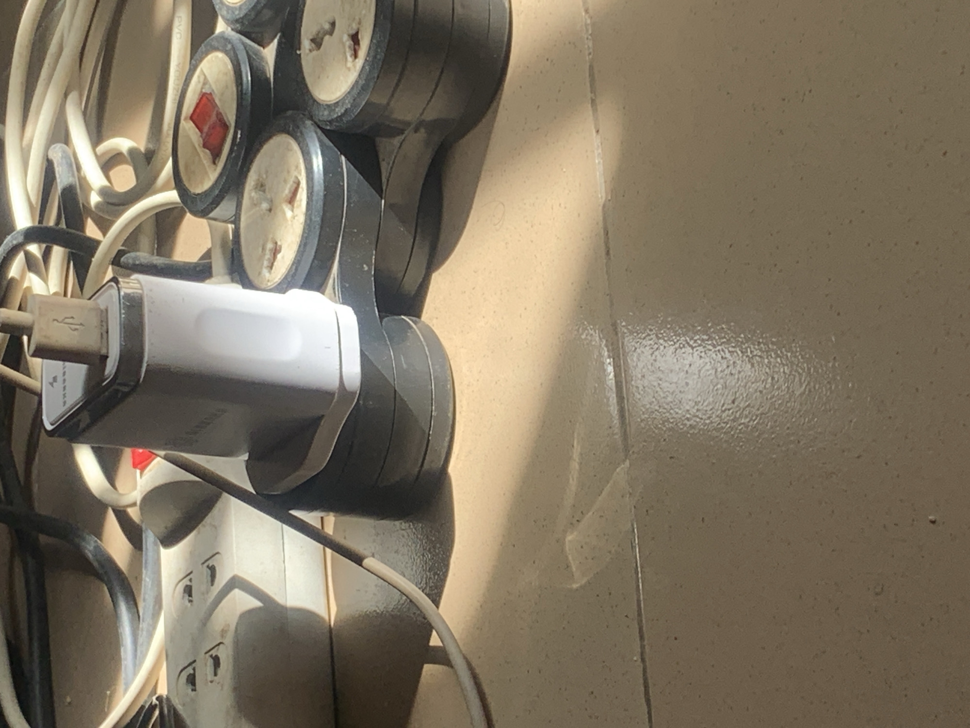 The image shows a close-up of a white Samsung phone charger plugged into a multi-outlet power strip. The power strip is connected to various cables, some of which are coiled around it. The scene appears to be indoors, with the objects resting on a light brown tiled floor. Sunlight is casting shadows on the floor and the objects, indicating the presence of a light source from the right side of the image. The overall impression is one of a cluttered electrical setup. There are no people or discernible landmarks that would place this scene specifically in Jauro Yinu, Nigeria.