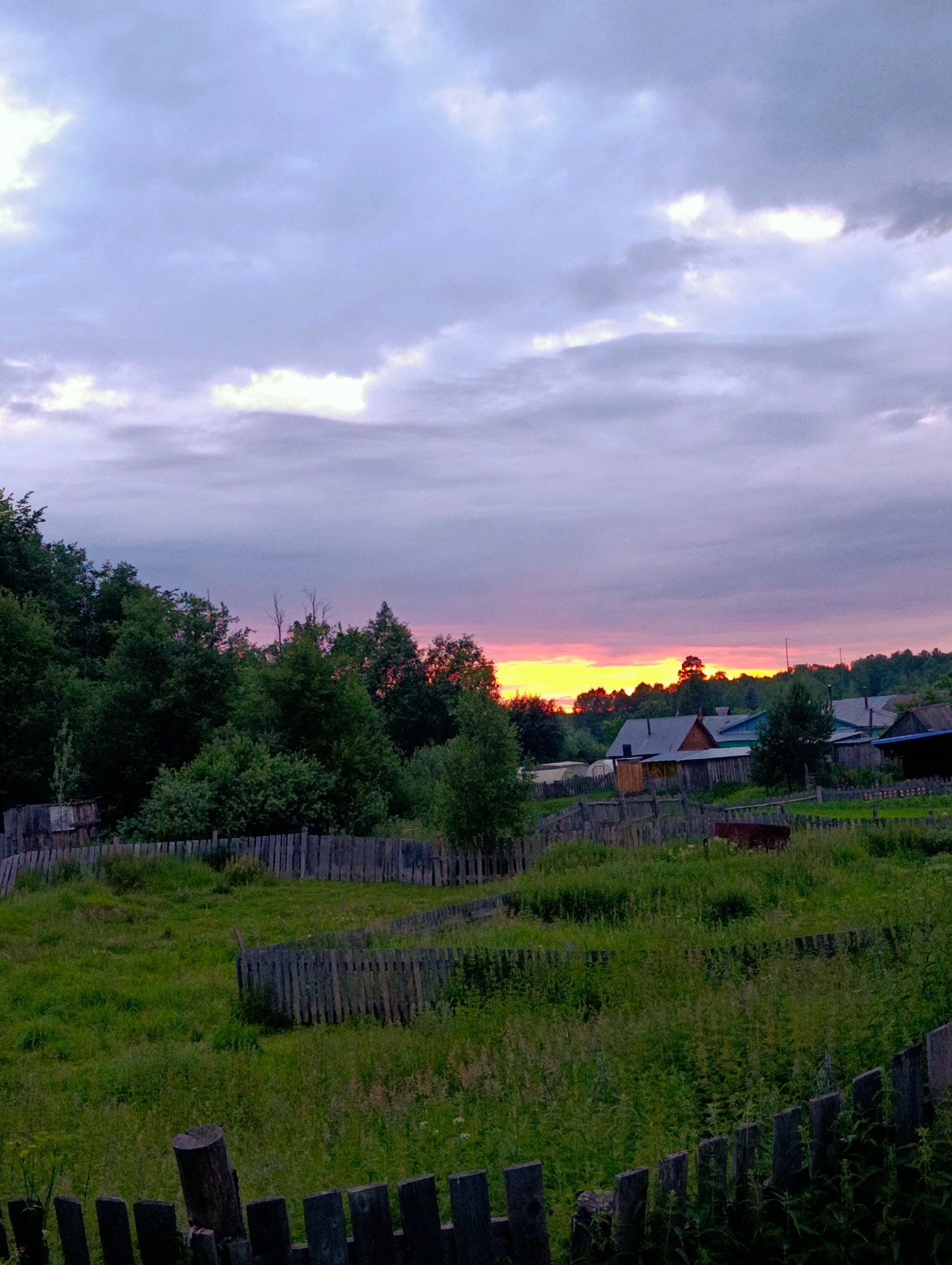 The image depicts a rural landscape in Буренка, Russia, during what appears to be sunset. The sky is filled with dramatic clouds, with vibrant oranges and yellows from the setting sun peeking through the darker, purplish-grey clouds. The light creates a serene and peaceful atmosphere.
In the foreground, a weathered wooden fence runs through a field of tall, green grass and weeds. Beyond the fence, the landscape transitions to a dense line of trees, primarily green foliage, with some bare branches visible. Further in the distance, a small village is nestled among the trees. Several wooden houses with dark roofs are visible, suggesting a traditional rural settlement.
The scene is tranquil, with no people or animals immediately apparent. The time of day is clearly sunset, indicated by the colors in the sky and the soft lighting across the landscape. The overall impression is one of quiet rural life at the end of the day, with a touch of natural beauty from the colorful sky. There is no visible text in the image.