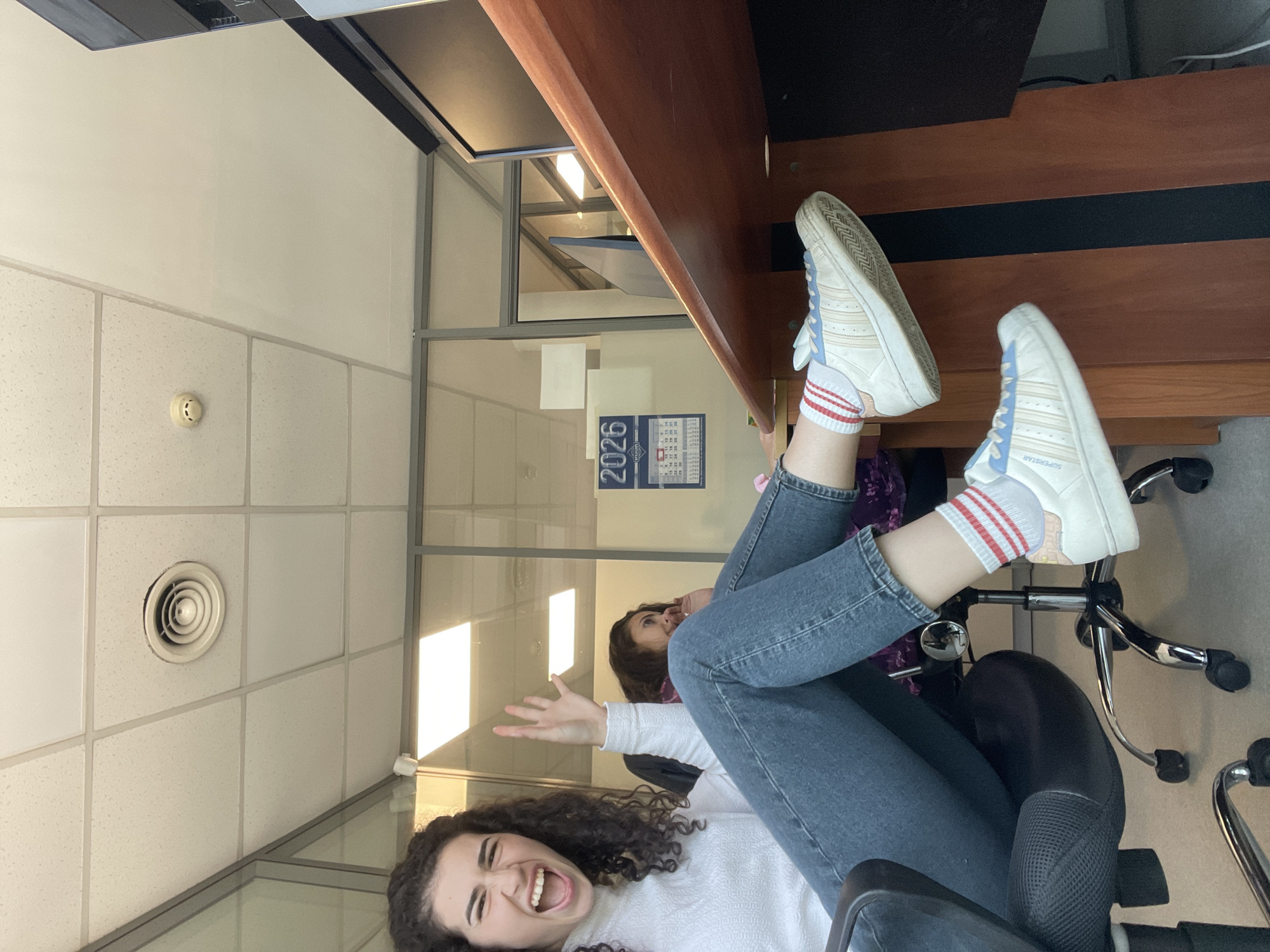 An indoor, vertically oriented photograph captures a playful moment in what appears to be an office or educational setting in Sofia, Bulgaria. The primary subject, a young woman with curly dark hair, lies reclined on an office chair with her legs propped up on a wooden desk. She wears a white long-sleeved top, blue jeans, white Adidas Superstar sneakers with light blue accents, and white socks with red stripes. Her face is turned towards the camera, mouth wide open in a joyful laugh, and one hand is raised in an open-palm gesture, conveying a sense of fun and amusement.
Behind her, partially visible, another person with dark hair looks upwards. The background features typical office elements: a drop ceiling with white acoustic tiles, a circular air vent, and a smoke detector. Glass and metal framed partitions divide the space, revealing bright light sources and a wall calendar for the year "2026." The desk is made of wood with a dark top section. The overall lighting is bright, suggesting daytime. The scene depicts a casual and lighthearted interaction, possibly during a break.