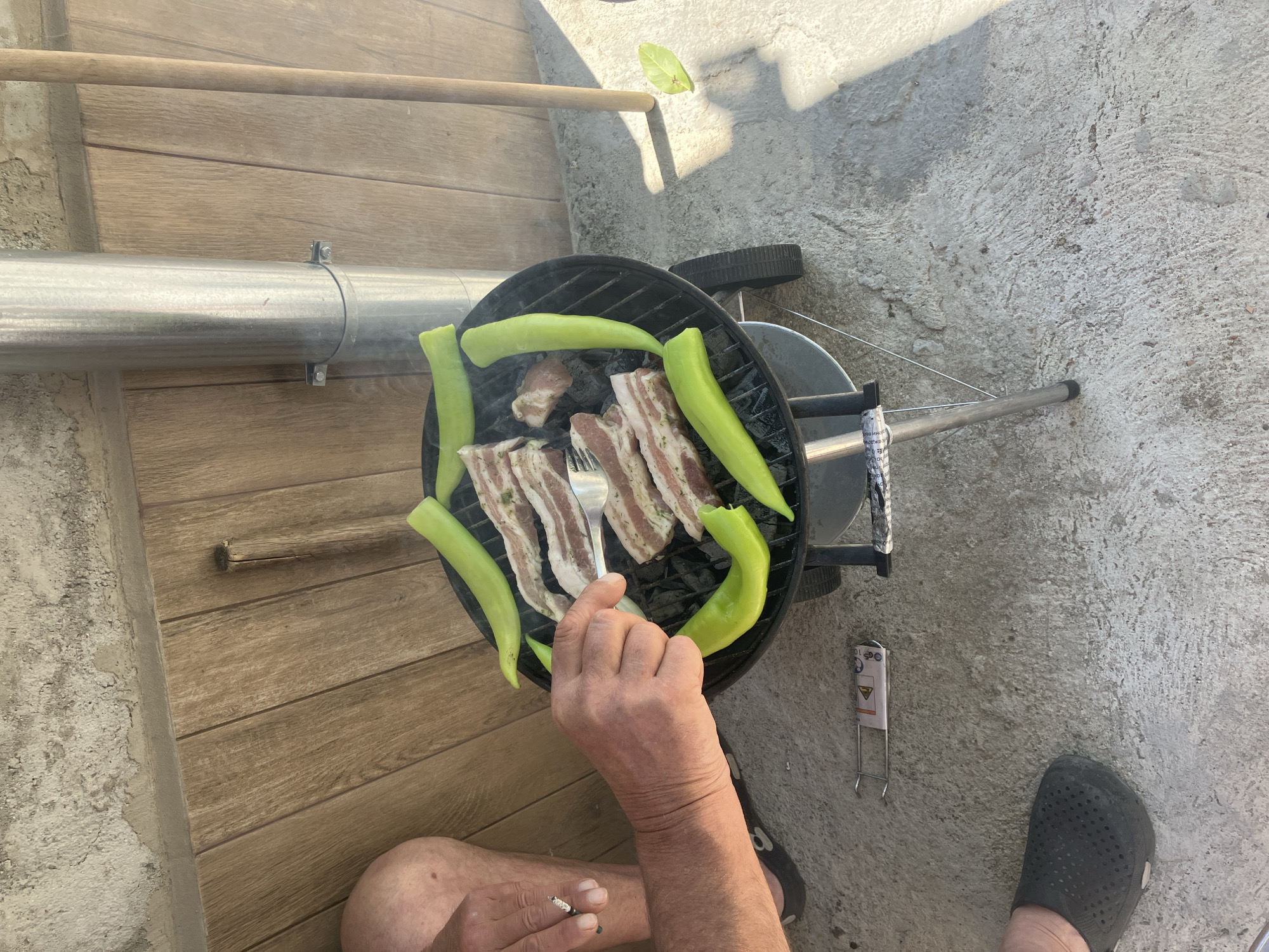 The image shows a person grilling bacon and green peppers on a small charcoal grill. The person is holding a fork and appears to be turning the bacon. The grill is located on a concrete surface next to a wooden deck. The person is wearing a black shoe. There is a cigarette between the person's fingers and a warning label laying on the concrete ground. The location is indicated as Komuniga, Bulgaria, though no elements in the image definitively confirm the location.