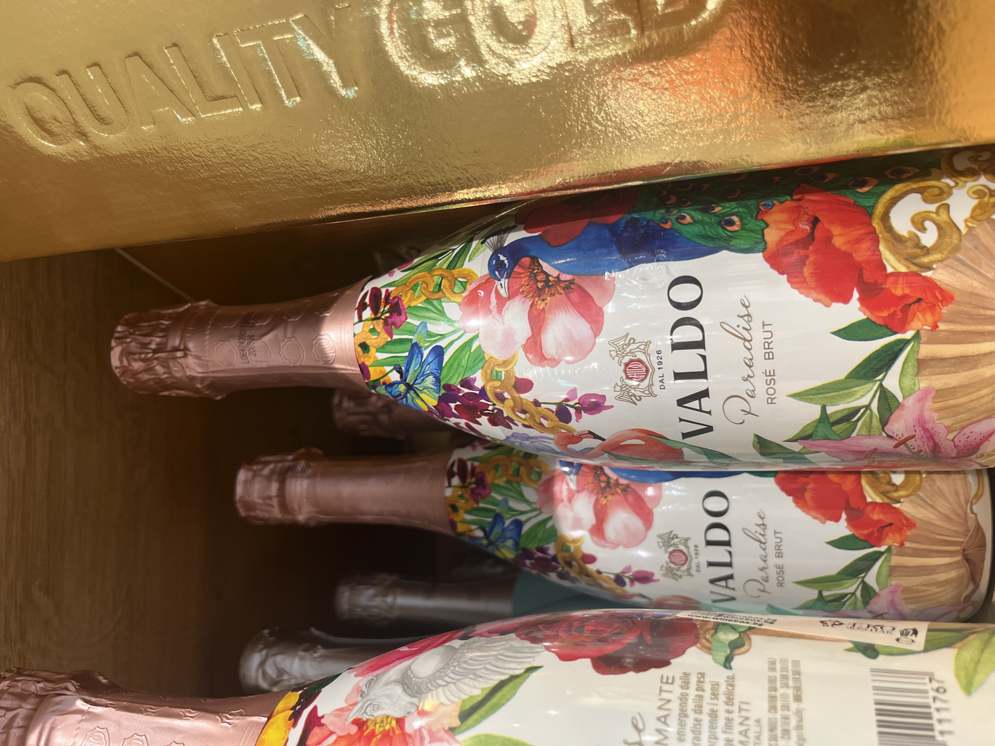 The image shows a close-up view inside a box labeled "QUALITY GOLD" containing bottles of "VALDO Paradise Rosé Brut" sparkling wine. The bottles are decorated with floral and peacock designs. Some text, including "DAL 1926", "Paradise Rosé Brut", and a bar code is visible on the bottles. There is no information to suggest the location is in Varna, Bulgaria, or anywhere else specifically.