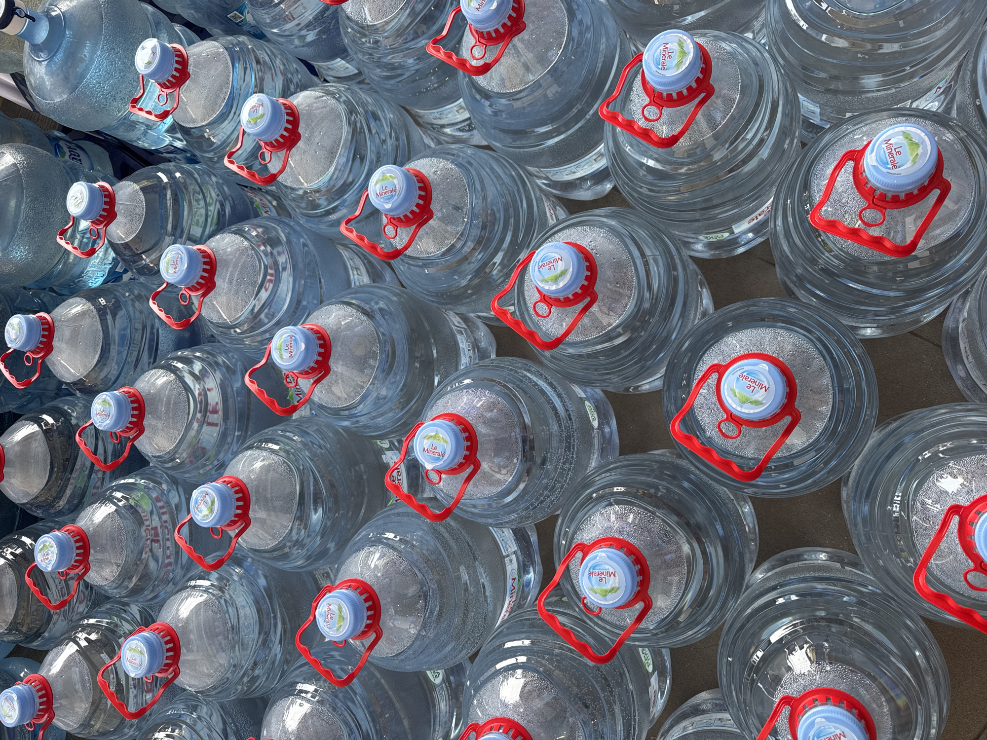 The image shows a collection of numerous large, transparent plastic bottles filled with water. The bottles are arranged closely together, creating a dense pattern that fills the frame. Each bottle has a distinctive blue cap with a red handle, and a label with the words "Le Minerale" on it. Based on the label, the location is likely in or near the city of Kuta Selatan, Indonesia, where this brand of mineral water is produced. The environment appears to be an indoor storage or display area, evidenced by the flat, neutral surface beneath the bottles. The lighting in the image is bright, casting gentle highlights on the curved surfaces of the bottles and the water inside. No people are visible.