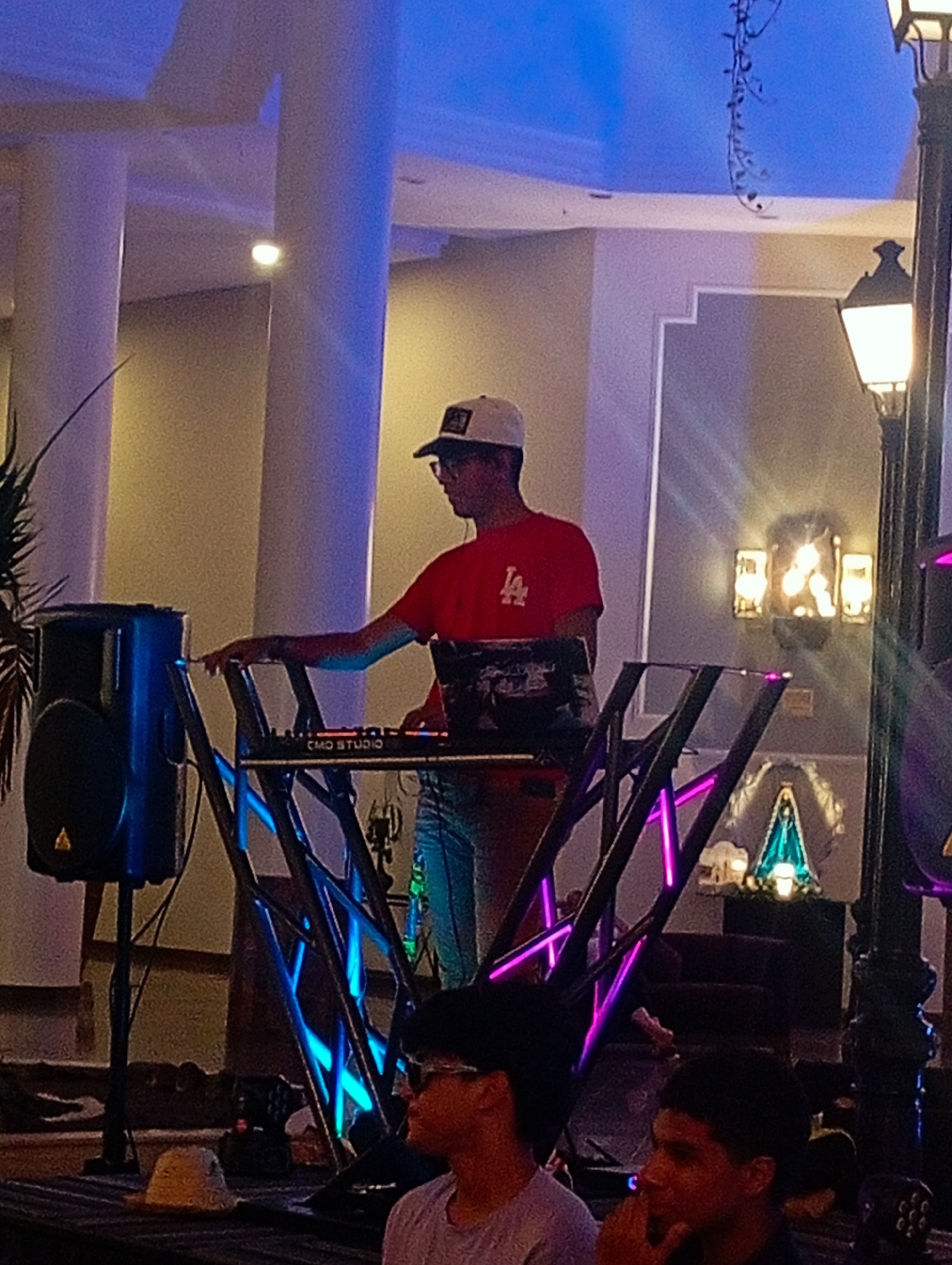 The image shows a DJ performing at an event, likely a party or gathering. The DJ, wearing a red t-shirt with an "LA" logo, a baseball cap, and glasses, is operating a "CMD Studio" mixing console on a table with a metal frame illuminated with blue and pink lights. A large speaker is positioned to his left. In the foreground, two individuals are seated, observing the performance. Behind the DJ setup, a building interior is visible, including columns, wall decorations, and lighting fixtures, including an illuminated religious icon. Based on the architectural style and known venues, this image could have been taken in Pedro González, Venezuela. A street lamp is partially visible on the right side.