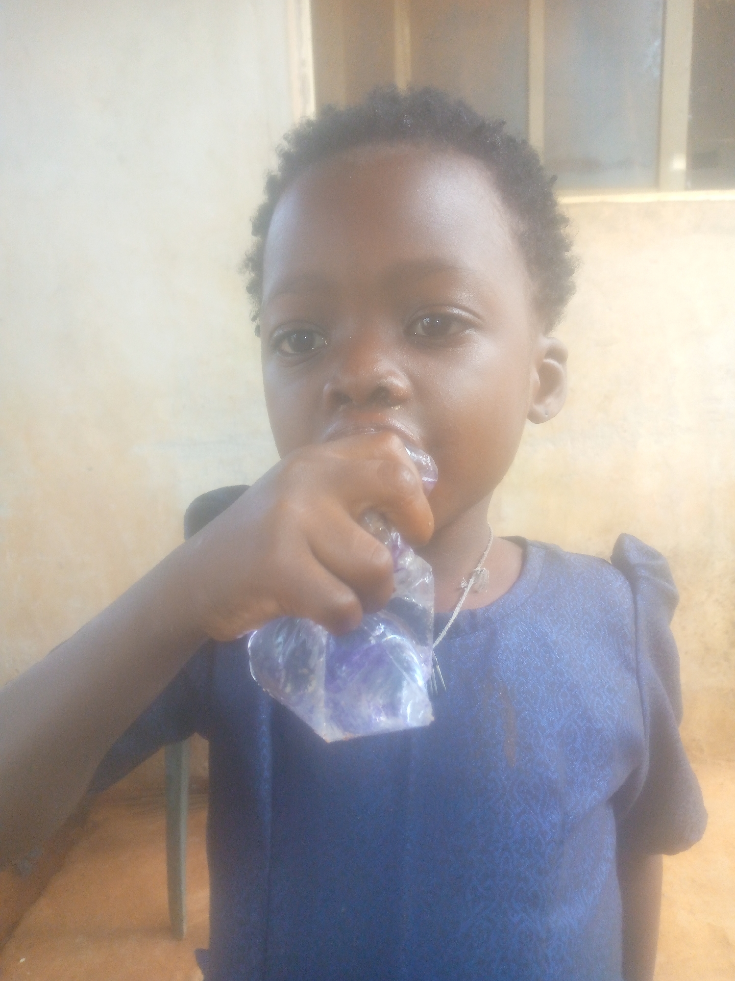 A young child with dark skin and short, curly dark hair is positioned centrally in a full-frame, eye-level shot. The child appears to be consuming something from a clear plastic sachet, which contains a purple substance, possibly a drink or ice. Their right hand holds the sachet to their mouth. The child is wearing a dark blue garment, likely a top or dress, with a subtle textured pattern. A thin, light-colored necklace with a small pendant is visible around their neck. The child looks directly towards the camera with a neutral expression.
The background consists of light-colored, plain walls, which appear to be either beige or light tan. The lighting is bright, causing some overexposure, particularly on the child's skin and the background walls, suggesting a well-lit environment, possibly indoors or in a sheltered area during the day. Part of a light brown or tan floor is visible at the bottom of the frame, along with a partial view of what appears to be a light green or blue chair leg to the left of the child. There is no visible text in the image.