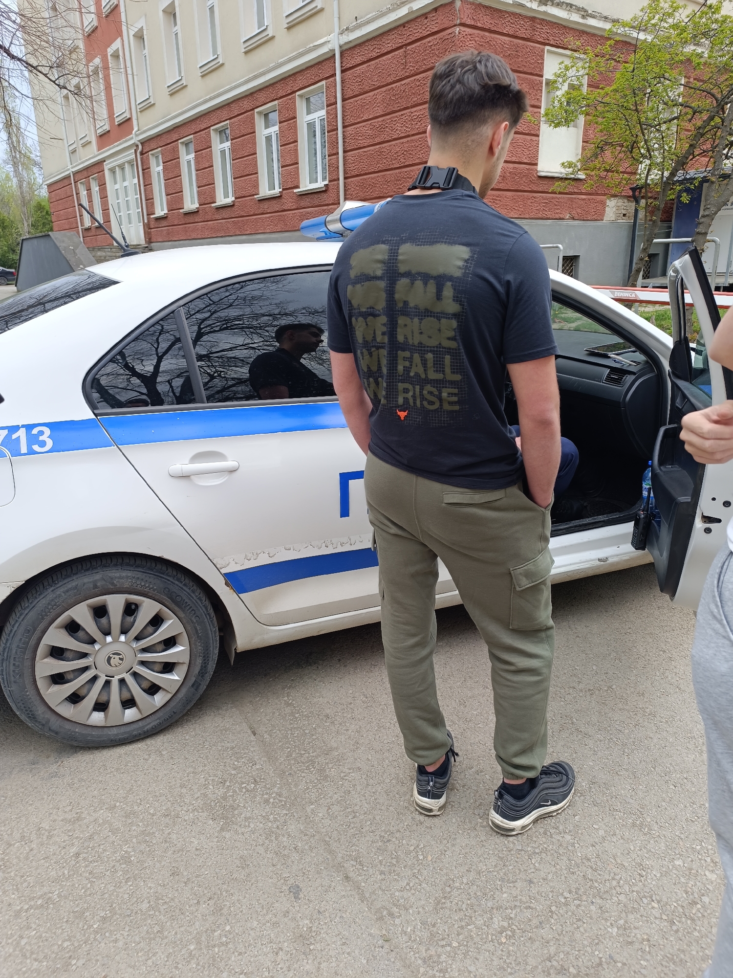 The image captures an outdoor daytime scene in what appears to be Sofia, Bulgaria. The central subject is a man viewed from behind, standing beside the open rear door of a white police car. The car features a distinctive blue stripe along its side, a blue emergency light on the roof, and partial Cyrillic text including "13" and "Г" (likely part of "ПОЛИЦИЯ" meaning Police).
The man is dressed in a dark t-shirt with the phrase "WE FALL WE RISE WE FALL WE RISE" printed on the back, accompanied by a small orange bull or horn logo. He wears olive green cargo pants and dark sneakers, with his hands in his pockets. A black strap or device is visible around his neck. Partially visible on the far right is another person's arm and side.
Reflected in the car's tinted rear window is the profile of a man, possibly a police officer or driver. Inside the car, a tablet is visible on the dashboard, and a water bottle along with a radio or similar device can be seen on the floor by the open door. The car's tire bears the brand "KUMHO" and "GROWING," with size "185/60 R15".
In the background, there's a multi-story building with reddish-brown textured lower walls and lighter upper walls, featuring numerous white-framed windows. Sparse tree branches suggest it might be early spring. The ground is paved asphalt. A red barrier, partially visible in the wider context, has the brand "BENINCA" written on it. The scene suggests an interaction or stop involving law enforcement.