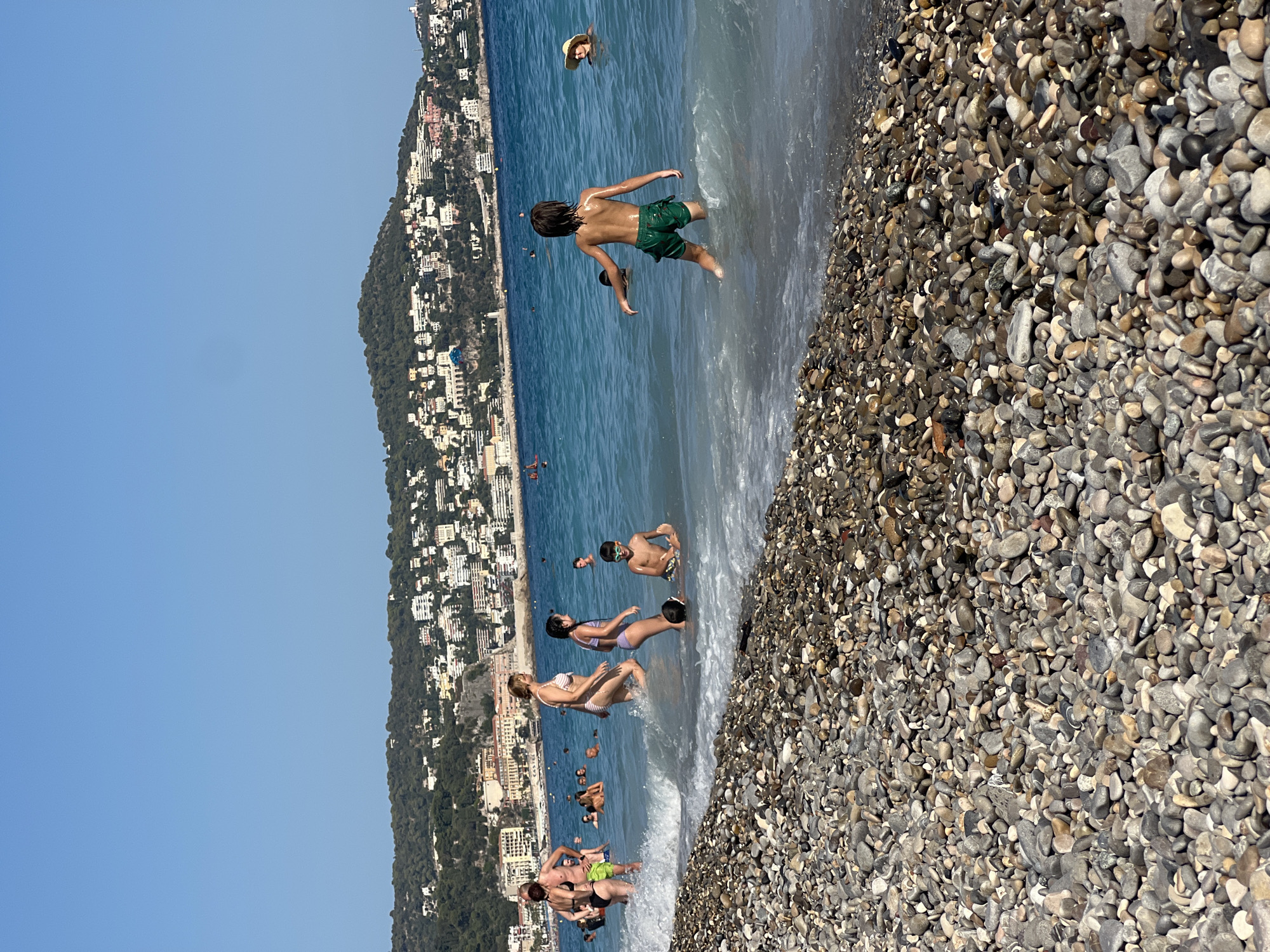 The image depicts a sunny day at a pebble beach in Nice, France, with the Mediterranean Sea in the background. A densely populated hillside town with white buildings and green trees overlooks the azure water. In the foreground, a person in green shorts stands in the shallow water, facing away from the camera. To the left, a woman in a striped bikini walks through the water, followed by two children, one in a bikini and another partially submerged. Further out in the sea, several other individuals are visible enjoying the water, including a woman wearing a straw hat. The beach itself is covered in smooth, multicolored pebbles.