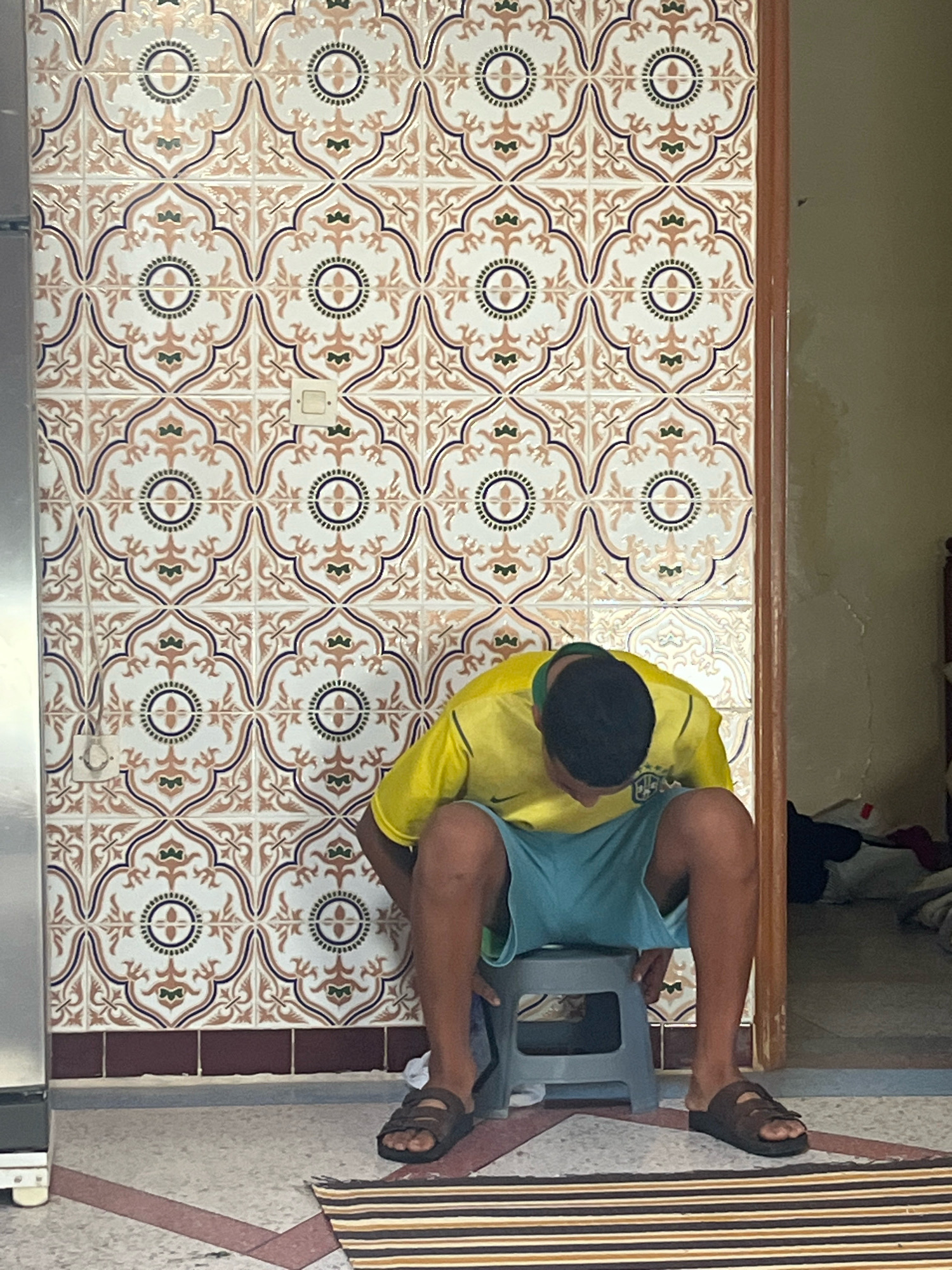 A young male wearing a yellow t-shirt and blue shorts is seated on a gray plastic stool. He is hunched over, facing downwards. He has dark hair and is wearing brown sandals. The background features a wall covered in patterned tiles with floral motifs in shades of white, orange, and dark blue. A light switch is visible on the tiled wall. To the right, a wooden door frame leads into a room with a bed and some clutter. To the left, a metallic appliance, possibly a refrigerator, is partially visible. The flooring is tiled with a geometric pattern, and a striped rug lies on the floor in front of the stool. This scene is located in the city of Azilal, Morocco.