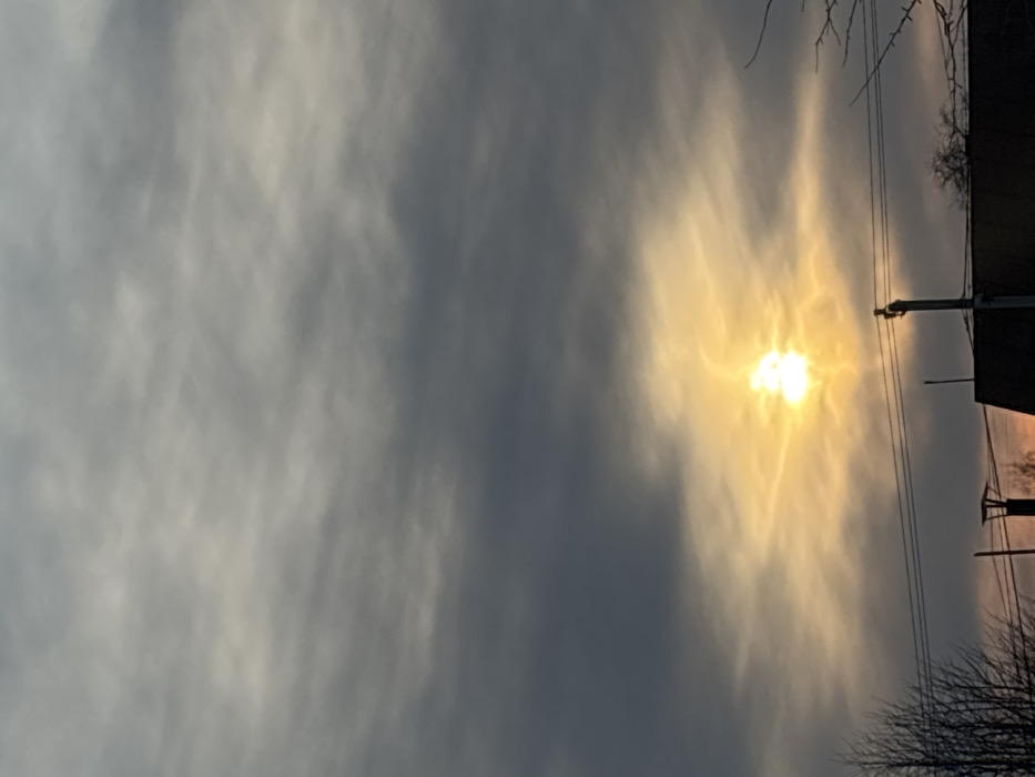 The image displays a cloudy twilight sky over Pavlovsky, Russia. A bright, intense yellow-orange sun is positioned in the lower-central portion of the frame, partially obscured by diffuse cloud cover, and emits distinct light rays. The sky transitions from dark gray, dense clouds in the upper two-thirds to lighter, more translucent clouds surrounding the sun. In the bottom fifth of the image, urban infrastructure is silhouetted against the luminous horizon. A dark utility pole with multiple horizontal electrical wires extends from the lower-center towards the right. To the lower-right, the dark, angular roofline of a building is visible, including a vertical chimney structure. Bare, leafless tree branches appear as dark silhouettes on the far left and right edges of the foreground. No human subjects are present.