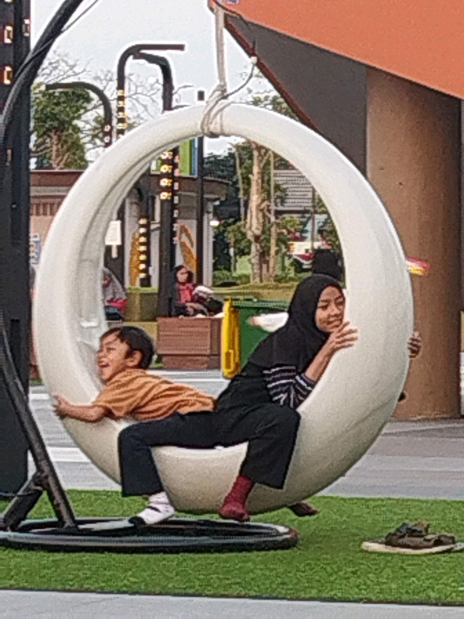This image depicts two individuals, a young boy and a young woman, seated within a large, egg-shaped hanging swing. The boy, wearing an orange t-shirt and dark pants, is positioned in front and facing left, with his mouth open in a wide smile. He is holding onto the swing. Behind him, the young woman, wearing a black hijab and a striped shirt with dark pants and red shoes, is also holding onto the swing. They are situated outdoors, on what appears to be a patch of artificial turf, with a grey paved area in the foreground. In the background, there are buildings, trees, and streetlights, suggesting an urban or commercial setting. The overall ambiance is one of leisure and enjoyment. The location is identified as Sidoarjo, Indonesia.