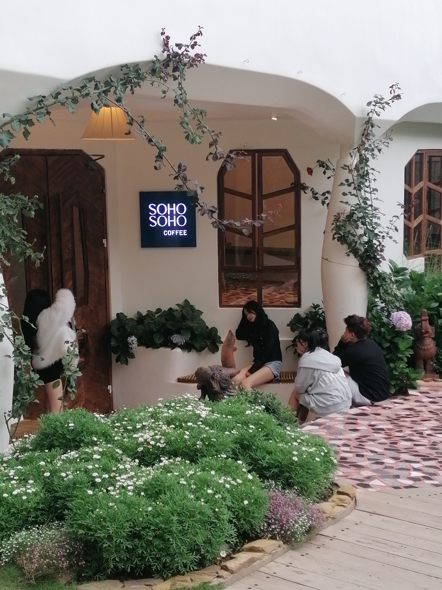 The image depicts the exterior of a cafe in Da Lat, Vietnam, likely named "SOHO COFFEE" as indicated by a illuminated sign. The cafe features a white, curved architectural design adorned with climbing vines. Several young adults are casually gathered outside. One individual, wearing a white fluffy coat and black clothing, stands near a wooden door. Three other individuals are seated on a low bench or ledge. One is wearing a black hoodie and denim shorts, another a grey hoodie and white pants, and the third a black t-shirt and dark pants. The foreground is filled with lush green bushes and white flowers, with a patterned tiled walkway visible to the right, leading to a wooden deck. The overall atmosphere suggests a relaxed, outdoor cafe setting.