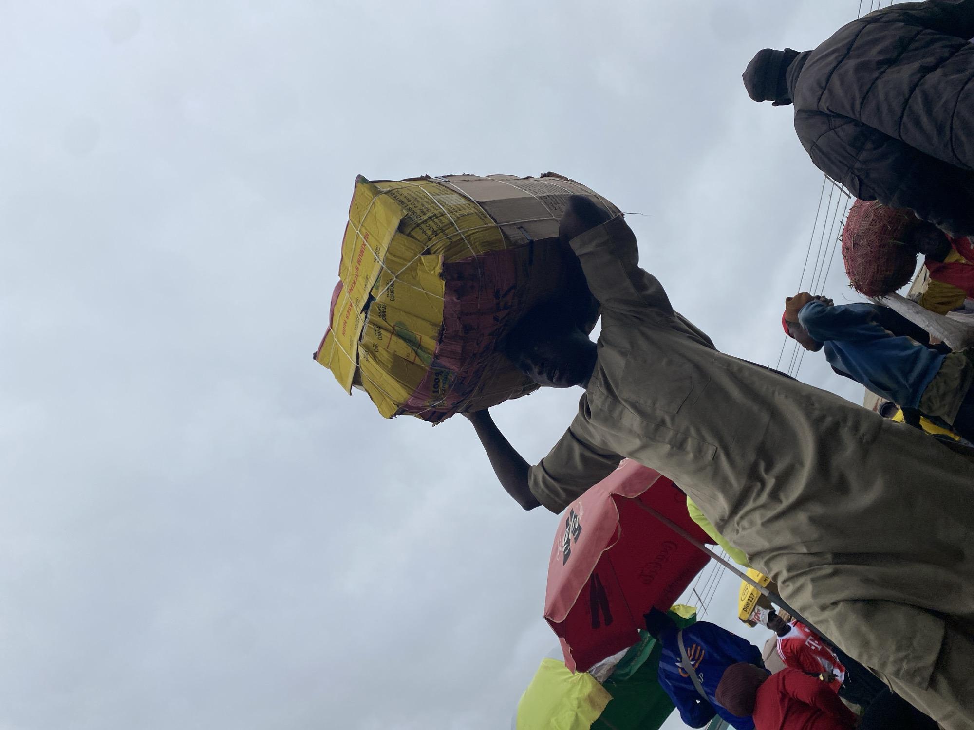 The image shows a man carrying a large, wrapped package on his head. He is wearing a tan, long-sleeved robe. The package is yellow with red and pink bands and appears to be secured with rope. The man is walking from a low angle perspective, with the sky as the background. Around him, several colorful umbrellas are visible, some red and some green, suggesting a market setting. Other people are also present, with some wearing jackets and hats. The overall scene depicts a busy outdoor environment, possibly in the city of Katon Rikkos, Nigeria, where people are engaged in trade or transport of goods. The sky is overcast, indicating a cloudy day.