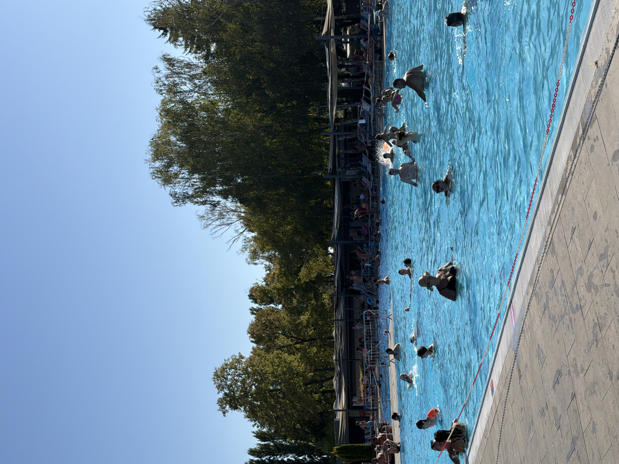The image depicts an outdoor swimming pool filled with people on a sunny day. The water is a vibrant blue, and the pool is surrounded by a paved area and lounge chairs under shaded awnings. Trees line the background, suggesting a park or recreational area. The sky is clear and blue. Numerous individuals are engaged in various activities within the pool, including swimming, wading, and socializing. Some people are standing in the water, while others are partially submerged or actively swimming. The overall atmosphere appears relaxed and enjoyable. This scene is consistent with a public swimming facility, such as the Thermal Park Dunajská Streda in Dunajská Streda, Slovakia, which is known for its large outdoor pools and recreational amenities.