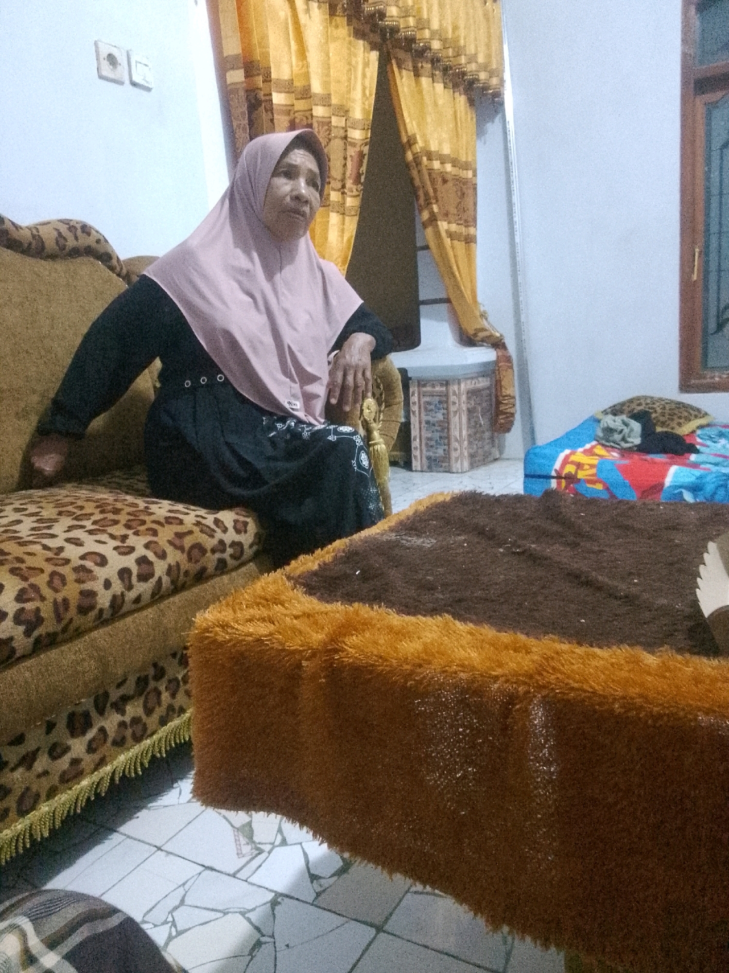 The image depicts an indoor scene, likely a living room, with elements suggesting a location in Krucil, Indonesia, due to the presence of a traditional patterned fabric and furniture styles common in the region. A woman wearing a light pink hijab and a dark abaya is seated on a sofa. The sofa has a leopard-print cushion and is adorned with gold fringe. In front of the sofa, a coffee table is covered with a brown shag rug and an orange fuzzy trim. To the right, a bed with colorful bedding is visible, along with another leopard-print pillow. Behind the woman, yellow and brown curtains are drawn, and a small decorative cabinet is situated against a white wall. Electrical outlets are visible on the wall near the curtains. The floor is tiled with a mosaic pattern. The overall impression is of a domestic, possibly humble, living space.