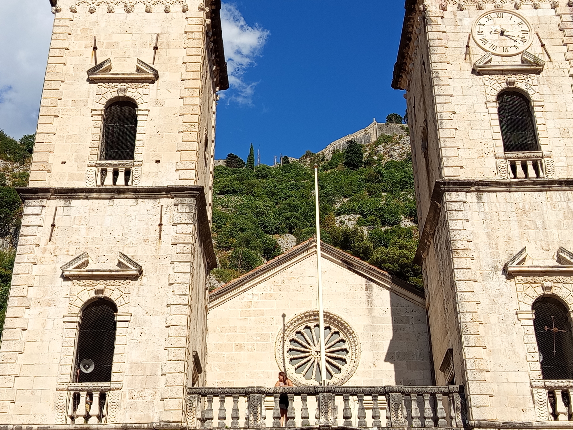 This media file captures a view of the St. Tryphon Cathedral in Kotor, Montenegro, under a bright, sunny sky. The scene is dominated by the cathedral's imposing stone facade, featuring two tall bell towers flanking a central section with a rose window. Roman numerals on a clock face are visible on the right tower. A person is standing on a balcony in front of the rose window, leaning on the railing and looking out. Lush green trees and a fortified wall on a steep hillside are visible in the background, suggesting the city's mountainous setting. The lighting indicates it is daytime, likely mid-morning or afternoon. The overall impression is one of historical grandeur and a peaceful, sunny day.