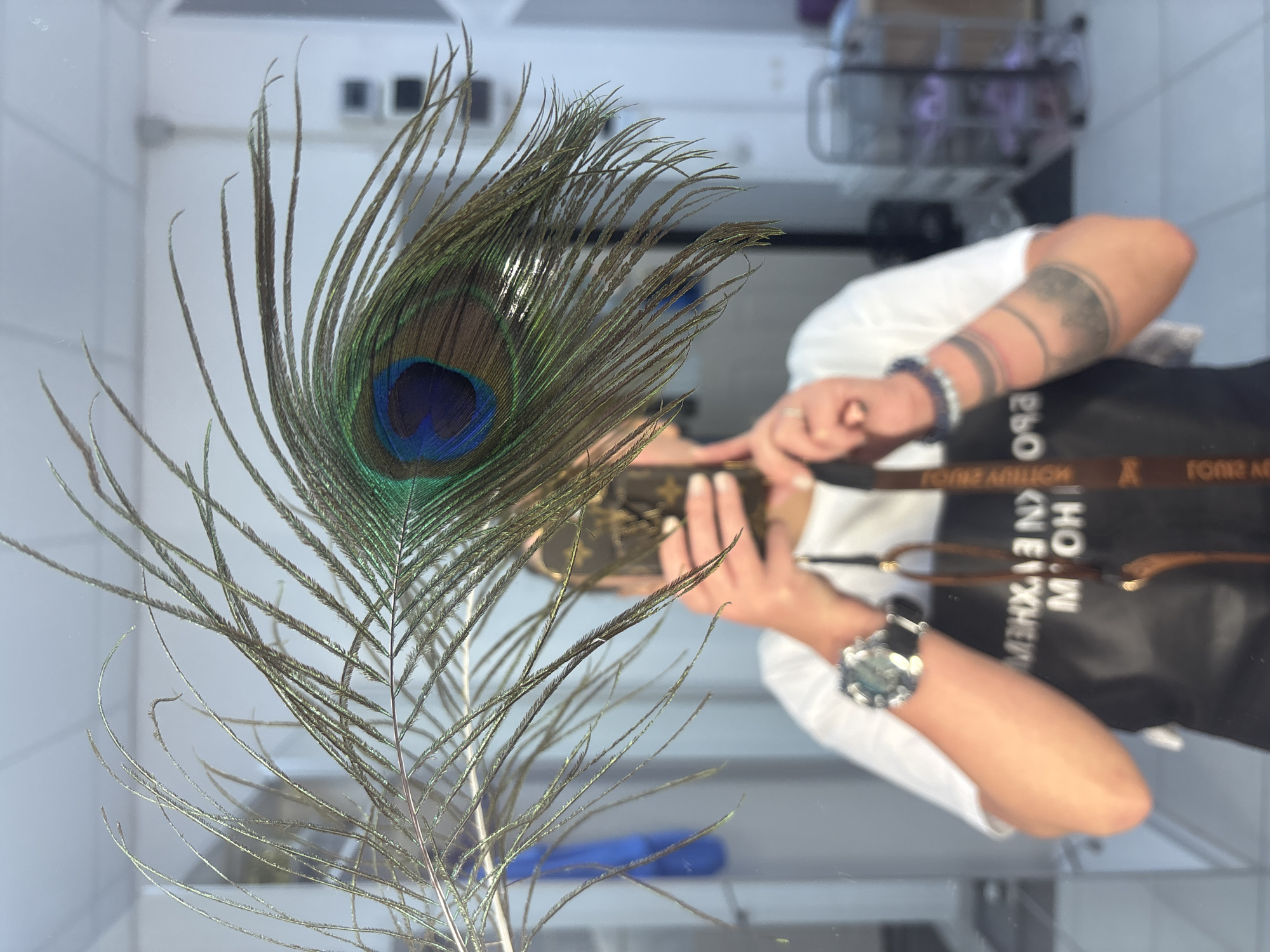 A peacock feather stands in the foreground. Behind the feather, the reflection of a person taking the photo is visible. The person is wearing a white shirt, a black apron with the word "HOME" and a brand that says "LOUIS VUITTON" visible on the strap. They are also wearing a watch and bracelets. In the background, blurred objects are present, possibly related to a retail environment. The location is potentially Perm, Russia.