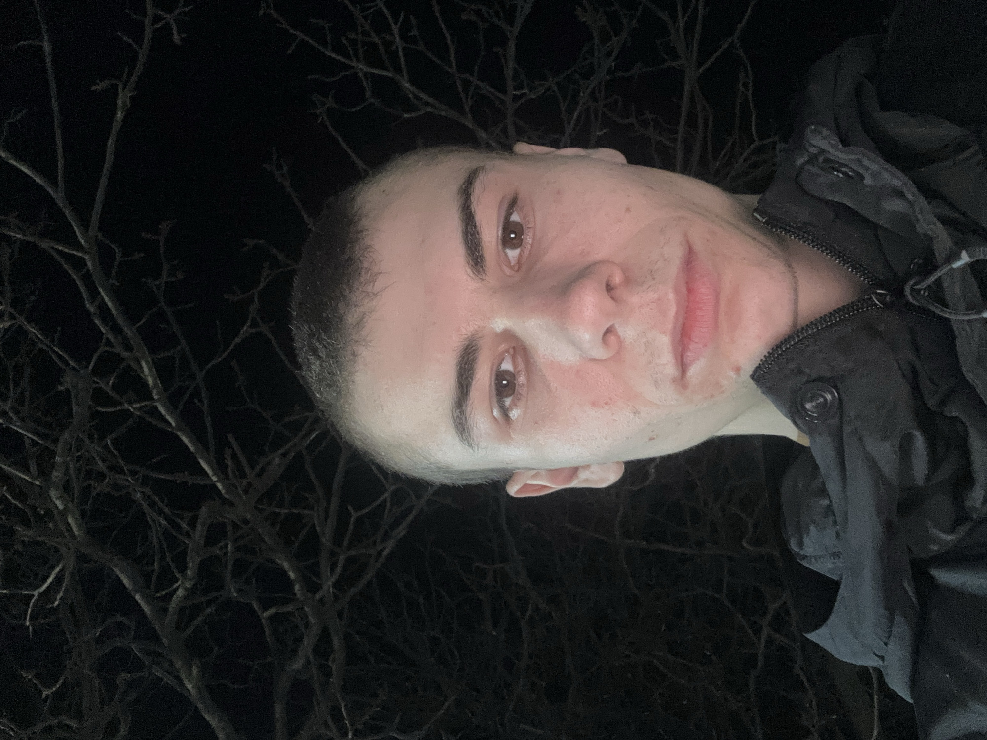 A head-and-shoulders portrait of a young man with short, dark hair and brown eyes is taken outdoors at night. The man is wearing a dark jacket with a zipper and buttons. Bare tree branches are visible behind him on the left side of the frame, set against a dark sky. The location cannot be identified as Plovdiv, Bulgaria, based on the provided information.