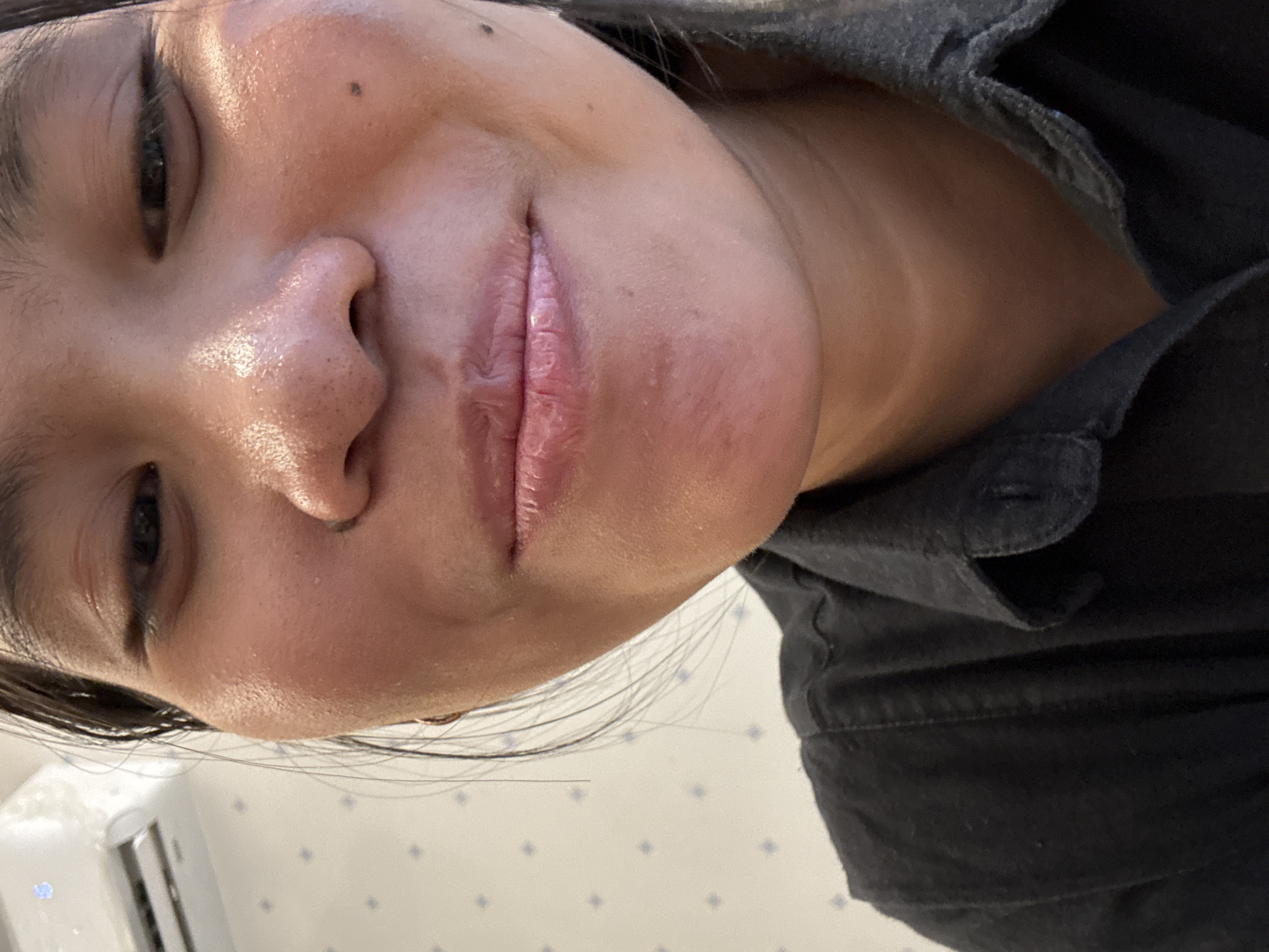 A close-up, over-the-shoulder shot captures the lower face of a young Indonesian woman in South Jakarta, Indonesia. She is wearing a black collared shirt, and her face is turned slightly to the left, with her lips parted in a subtle smile. Her skin appears slightly dewy, and a small mole is visible on her right cheek. In the background, a white air conditioning unit is mounted on a wall adorned with a light blue grid pattern. The lighting is bright, highlighting the woman's features and creating a slight sheen on her skin. The overall impression is of a casual, candid moment.