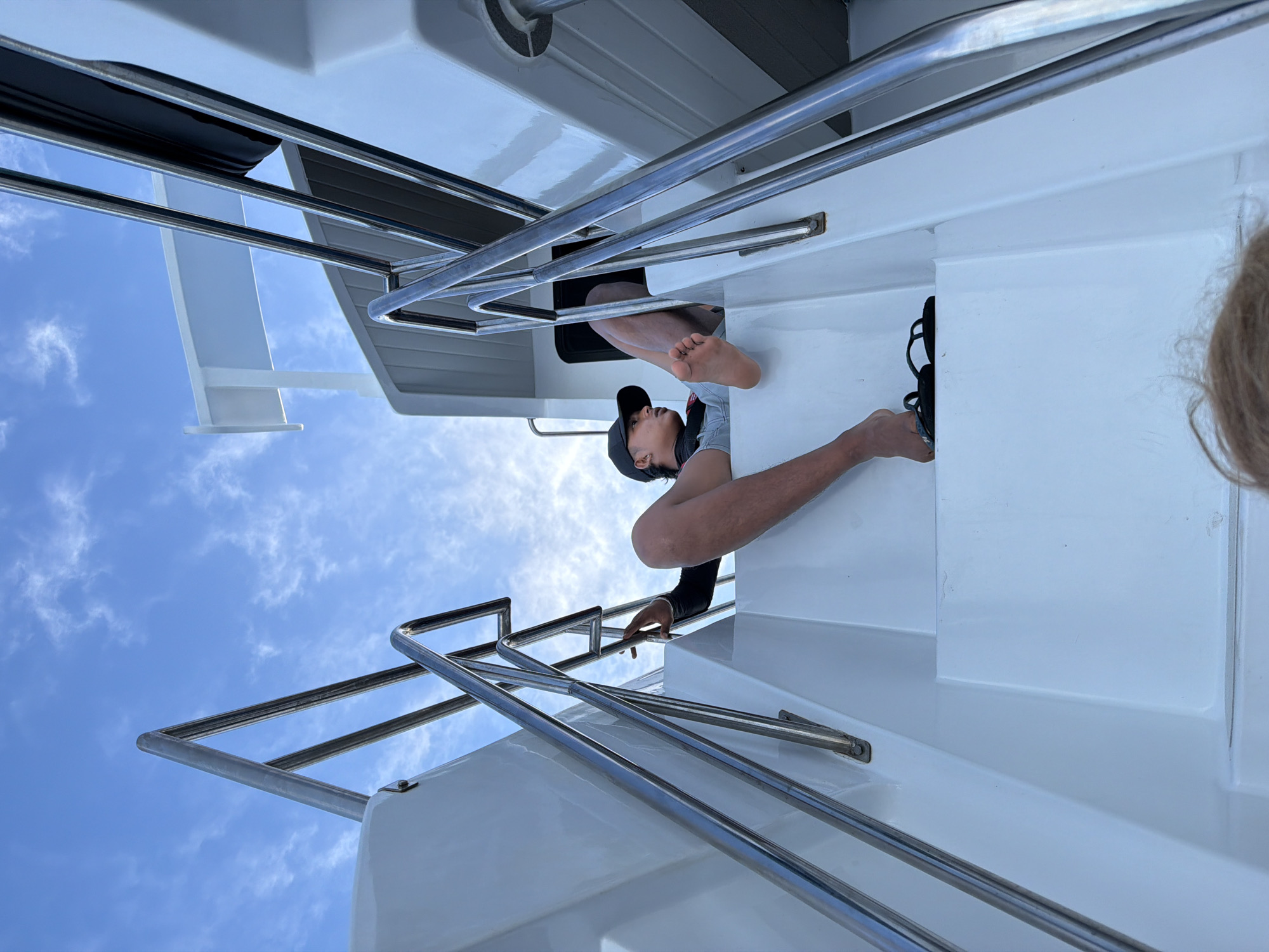 The image, originally rotated 90 degrees clockwise, depicts a scene on a white boat or yacht under a bright blue sky with scattered white clouds. The perspective is from a low angle, looking upwards at the vessel's structure.
The primary subject is a person, likely a young male, in the process of emerging or climbing through an opening or hatch in the boat's white deck. He is positioned centrally, wearing a dark baseball cap, a light-colored shirt, and a dark long-sleeved top. His bare feet and legs are visible as he navigates the tight space, with his right hand gripping a polished silver metal railing for support.
The boat's surfaces are predominantly white and appear clean and well-maintained. Several prominent silver metal railings run horizontally and at angles, indicating a staircase or access point between different levels of the boat. On the far right, a small part of another person's head, with blonde or light brown hair, is visible, and near their hand, black shoes or sandals are tucked into a recess.
The clear sky and strong lighting suggest it is daytime with pleasant weather. The scene captures a moment of activity or transition on the watercraft. There is no visible text in the media file.