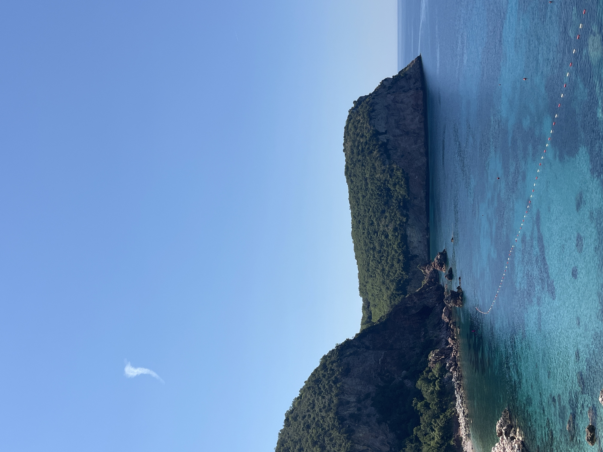 An aerial shot captures a serene coastal scene in Sveti Stefan, Montenegro, under a clear blue sky. The foreground is dominated by a tranquil sea with crystal-clear turquoise waters, revealing the sandy seabed below. A white and red buoys line demarcates a swimming area, with several red buoys floating further out. Rocky outcrops punctuate the shoreline, giving way to a secluded beach. Lush green hills rise steeply behind the coast, their slopes covered in dense vegetation. Two prominent headlands, one on the left and another on the right, frame the bay. A wispy white cloud, possibly a contrail from an aircraft, drifts lazily in the vast expanse of the sky.