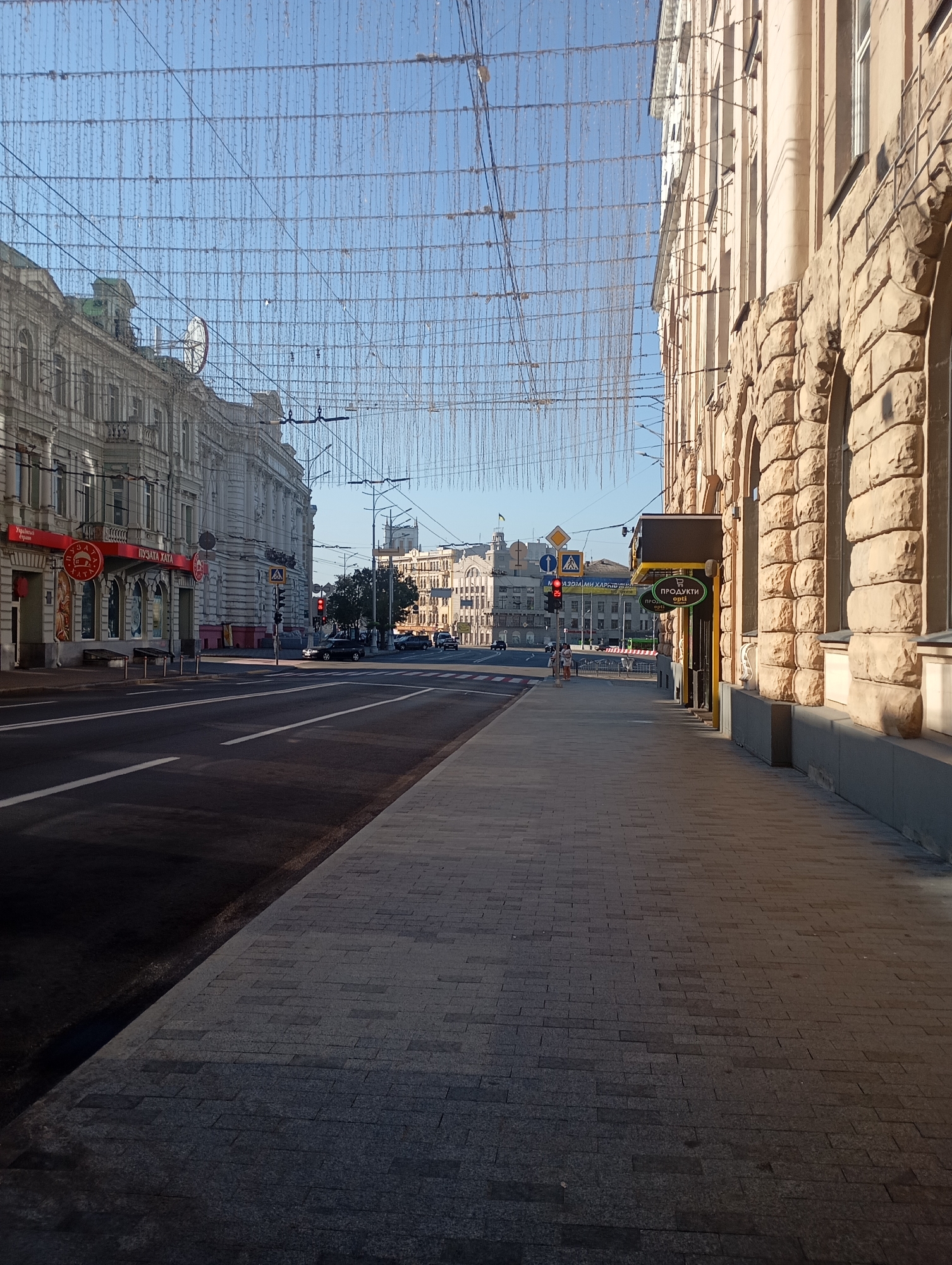 A street scene in Kharkiv, Ukraine, is depicted under a clear blue sky. The street is lined with old, ornate buildings on both sides. Decorative strings of lights hang overhead, crossing the width of the street. The building on the left has a classical facade with detailed architectural elements and signage for "ПУЗАТА ХАТА". The building on the right features a stone facade with arched windows and a sign that reads "ПРОДУКТИ". A paved sidewalk runs along the right side of the street, and the road itself is marked with white lines. Several cars are present on the road, and a traffic light is visible, showing red. A few pedestrians are walking on the sidewalk in the distance. The overall impression is a quiet urban street, possibly in the morning or late afternoon given the lighting.