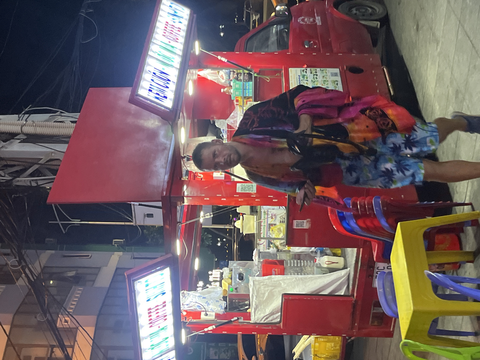 The image depicts a man standing in front of a red food truck at night in Bac Nha Trang Ward, Vietnam. The truck has signage in Vietnamese. The man is wearing patterned shorts and a colorful shirt. There are stacks of colorful plastic chairs near the truck. Power lines and buildings are visible in the background.