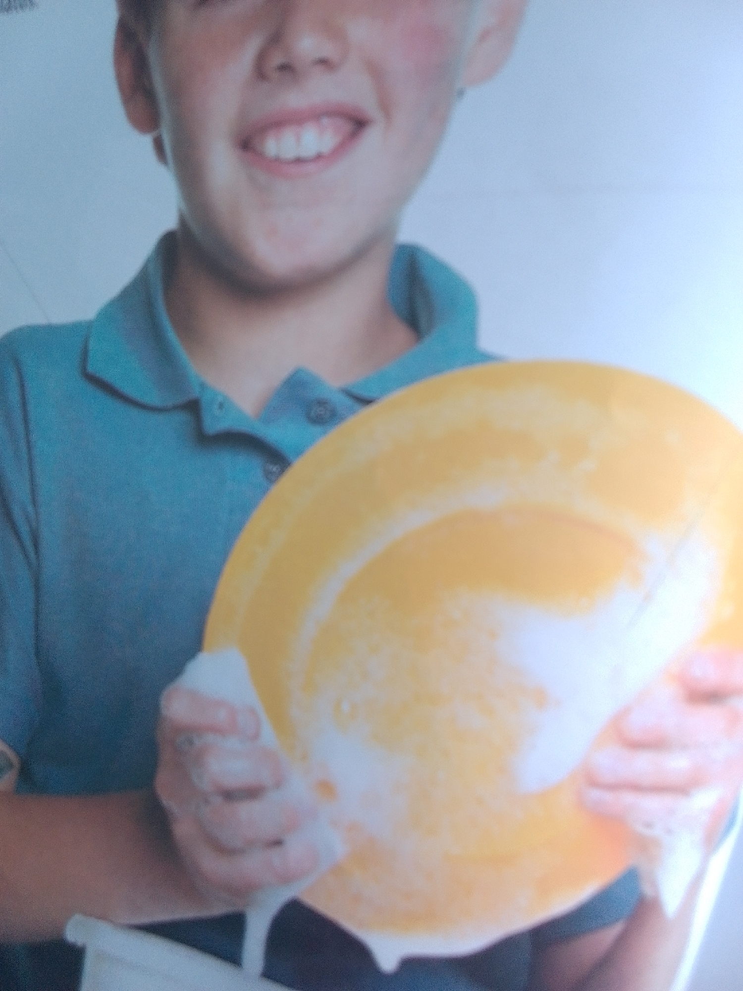 The image shows a close-up of a young boy, wearing a blue collared shirt, washing a yellow plate. He is holding the plate with both hands, and it is covered in white soap suds. The boy's face is visible, and he is smiling. The background is out of focus and appears to be a light blue or white wall. There is no indication of the location being La Tagua, Venezuela.