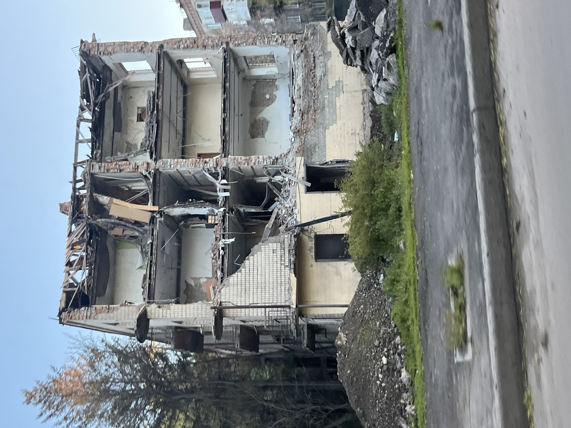 The image shows a severely damaged building in Kryvyi Rih, Ukraine. The building is missing large sections of its facade, exposing the interior structure. Debris is scattered around the base of the building and along the adjacent road. The building is multi-storied, with visible remnants of walls, floors, and ceilings. Other buildings are partially visible in the background. Trees and vegetation are present in the lower left corner of the image.