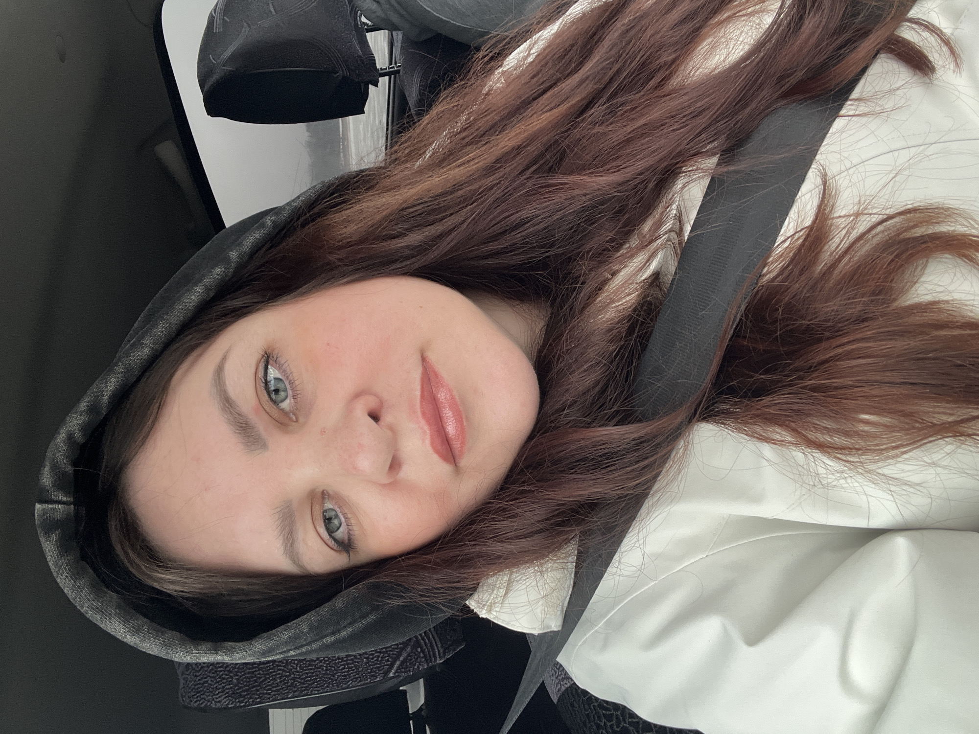 A woman with light skin and long brown hair is wearing a dark gray hoodie with the hood up and a white shirt or jacket. She is also wearing a black seatbelt across her body. She appears to be inside a car. The location in the city of Noviy, Russia, is not evident in this image.