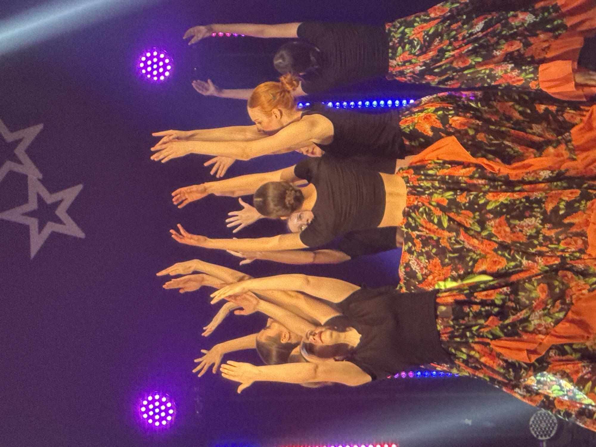 A group of five dancers is performing on a stage in Nizhny Novgorod, Russia. They are wearing black crop tops and long skirts with an orange floral pattern. The dancers are lined up in a row, with their arms extended forward. The stage is lit with purple and blue lights, and there are star-shaped decorations on the backdrop.