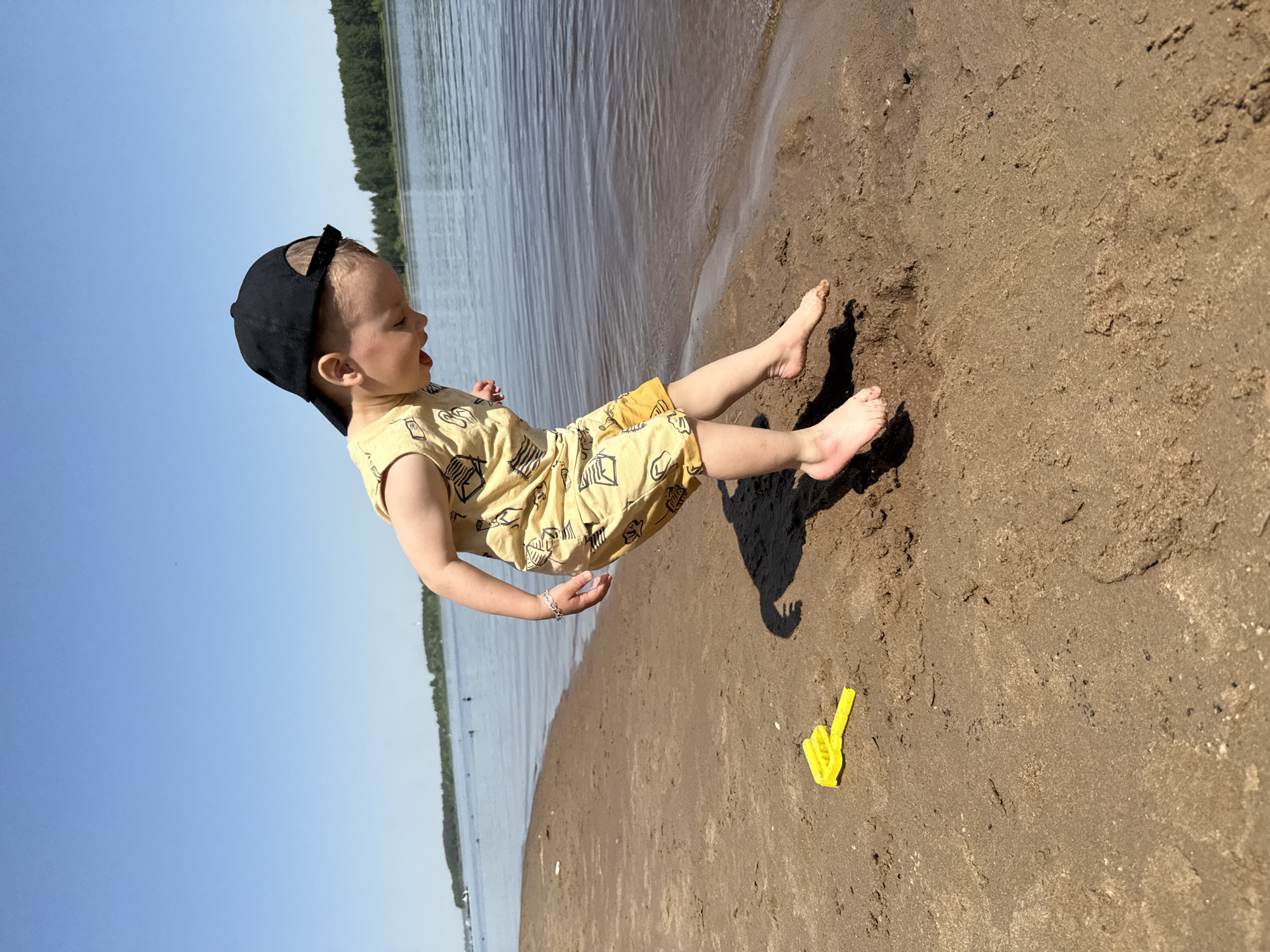 A young child is pictured playing on a sandy beach adjacent to a body of water. The child, wearing a light yellow outfit with a dark baseball cap, is in motion, with legs extended and feet off the ground. A yellow plastic toy is on the sand nearby. The background shows water and trees, suggesting a lakeside or coastal environment. The image may be taken in Rusanovo, Russia.