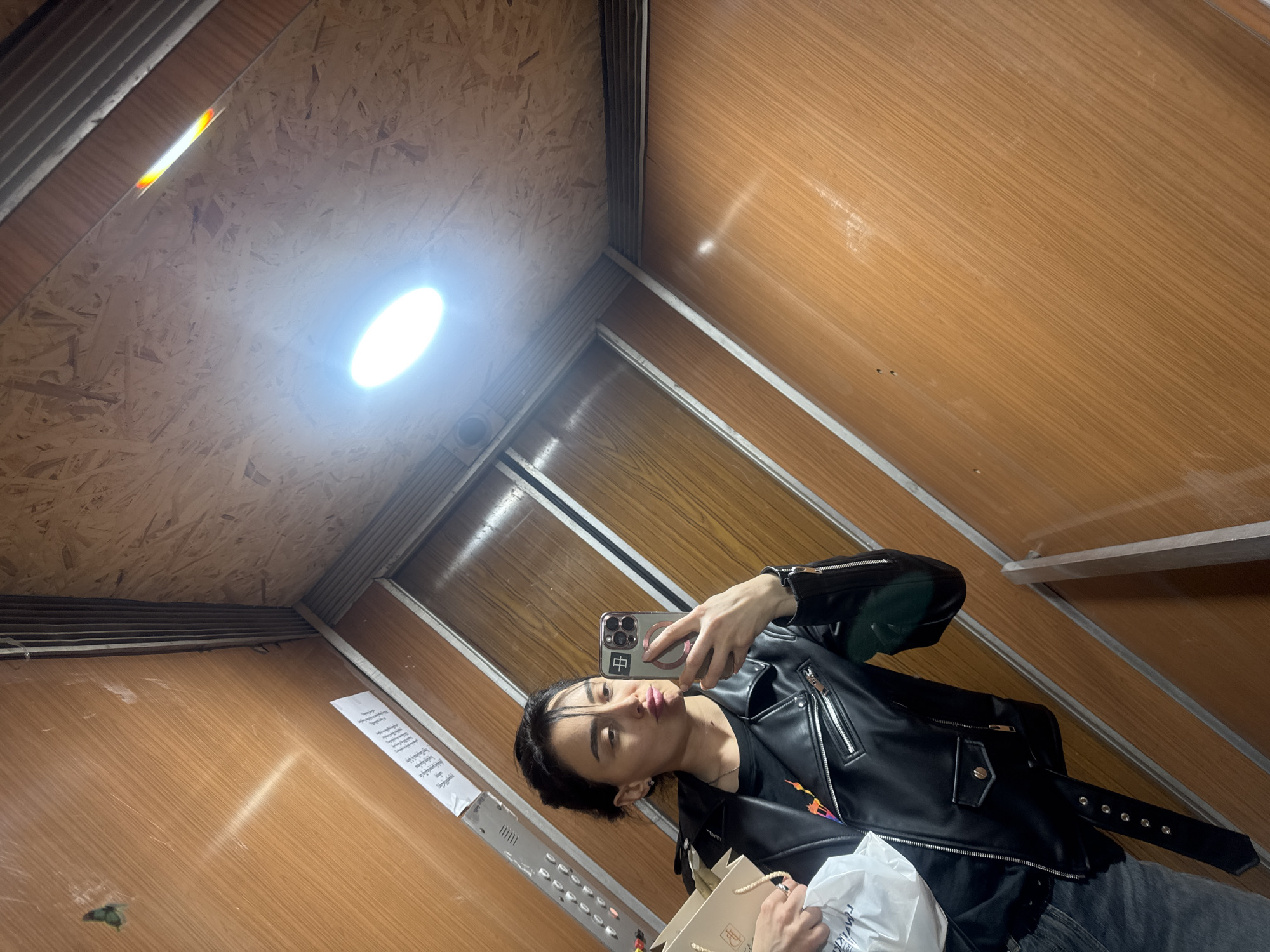 A young adult stands in a small elevator with wood-paneled walls and a plywood ceiling, illuminated by a central light. The individual holds a smartphone in their right hand, capturing a selfie. They wear a black leather jacket over a dark t-shirt and gray jeans, also holding a shopping bag in their left hand. The location, indicated by Georgian text on a sign, suggests the city of Tbilisi, Georgia. A panel of elevator buttons is visible on one wall.
