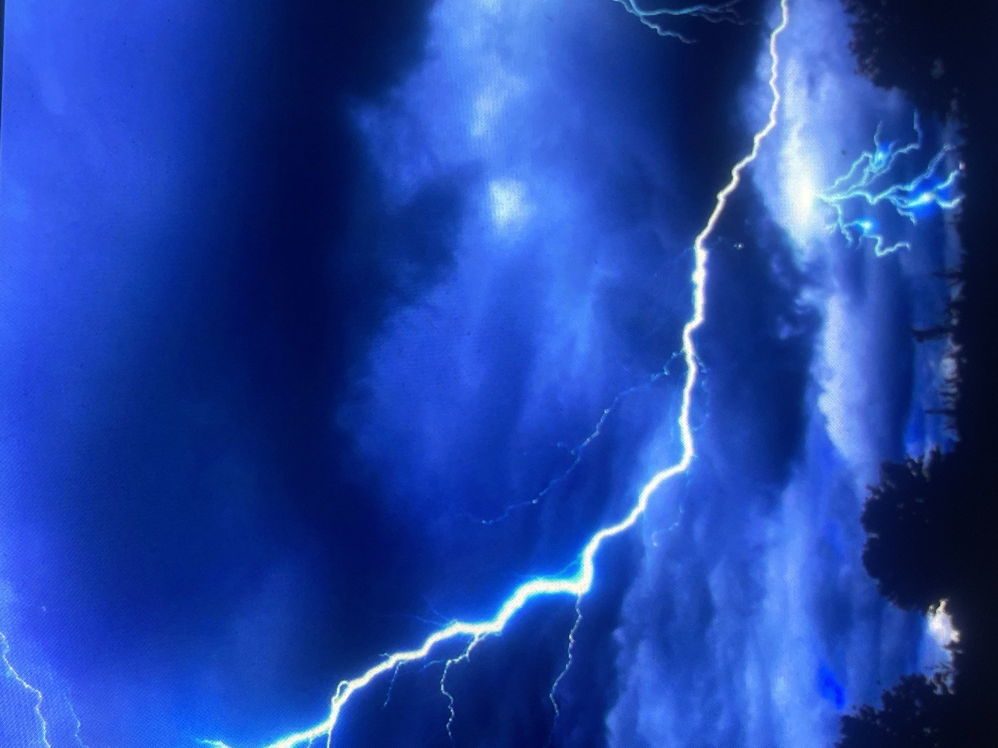 The image depicts a dramatic nighttime lightning storm set in the city of Zelenyi Hai, Ukraine. The sky is dominated by dark, heavy clouds rendered in various shades of deep indigo and electric blue. A prominent, intensely bright, electric-blue and white lightning bolt originates from the upper left, extending jaggedly across the mid-upper sky towards the right. A secondary area of bright blue-white illumination, featuring multiple branching lightning strikes, is visible in the lower right, illuminating a cloud formation from within. The horizon is defined by a dark, silhouetted line of dense tree canopies and low vegetation. The overall lighting is provided by the intense flashes of lightning, creating strong contrast against the dark environment. A fine, consistent grid pattern, characteristic of a screen display, is visible across the entire image, indicating it is a photograph of a digital image shown on a screen rather than a direct photograph of the natural event. No people or specific man-made structures are discernible.