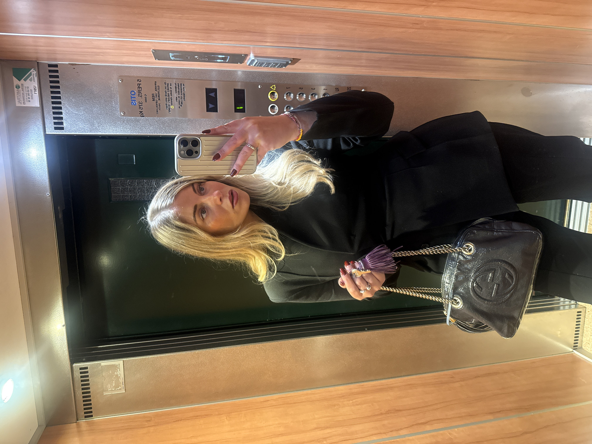 Here is a detailed description of the image:
**Scene:** The photo appears to be a selfie taken by a woman inside an elevator. The elevator has wood paneling on the walls and a metallic control panel with buttons for floors and a digital display.
**People:** The main subject is a woman with blonde hair. She is holding a smartphone, presumably taking the photo. She is wearing a black jacket or blazer.
**Objects:** Other notable objects include a black Gucci purse with a chain strap and a tassel. The woman is also wearing jewelry, including rings and a bracelet. The smartphone is visible, showing a metallic back.
**Actions/Interactions:** The woman is taking a selfie, which involves holding the phone out in front of her.
**Environment:** The environment is an elevator with wood-paneled walls and a metal control panel. The lighting appears to be artificial.
Based on the text "SIMPRA" on the control panel, the elevator is likely a SIMPRA brand elevator. The elevator control panel also has text in a language that appears to be either Greek or Russian, but it can't be clearly defined from the OCR.
**Location:** It is not possible to determine from the image if the location is Paris, France, or anywhere else for that matter.