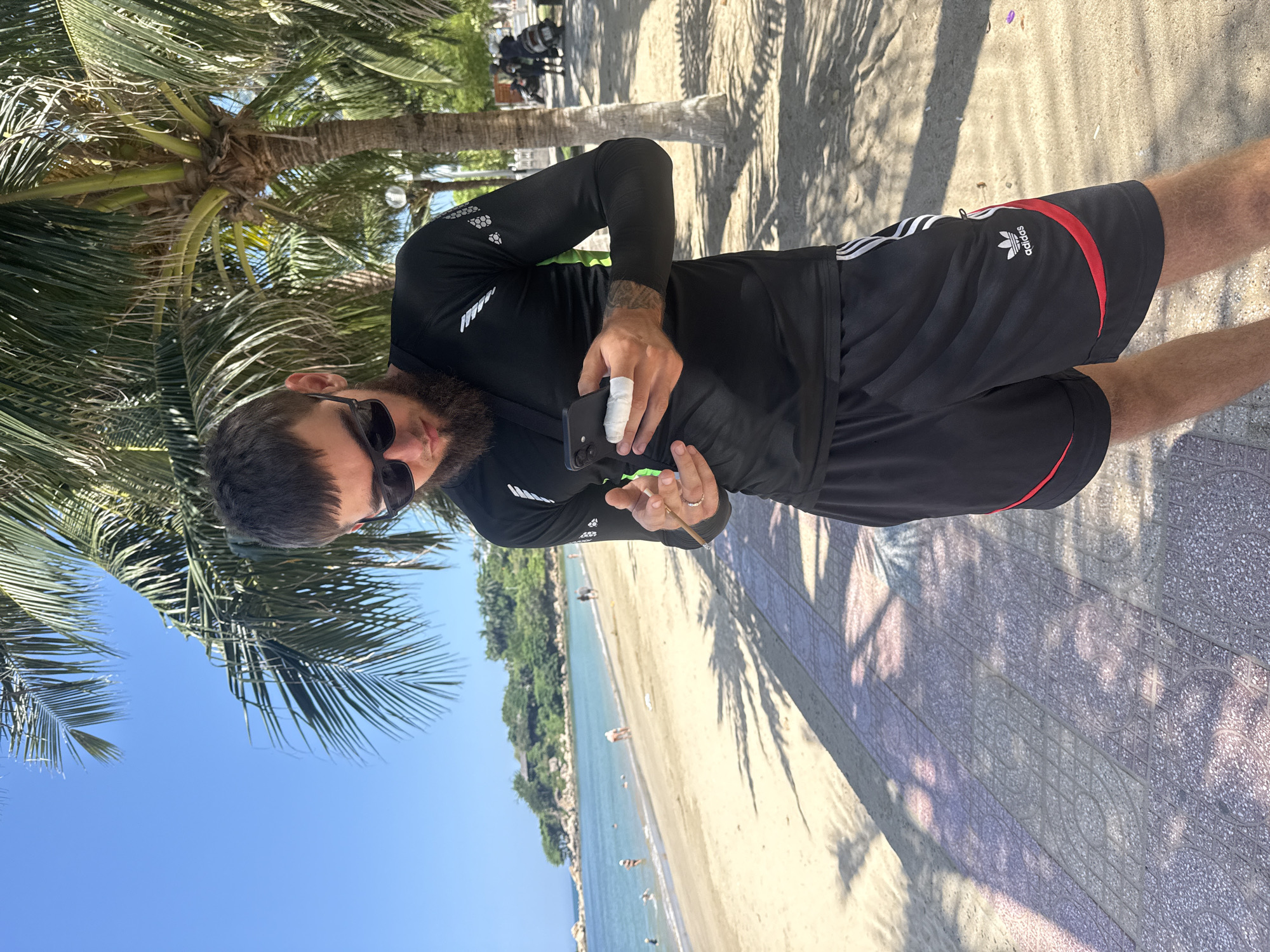 A man with a beard and sunglasses stands on a paved walkway next to a beach. He is wearing a black long-sleeved rash guard with green accents and black Adidas shorts with red trim. He is looking down at his phone, which he holds in his right hand. His left hand is visible near his phone, and he appears to be touching it. On his left thumb, there is a white bandage.
In the background, there is a sandy beach with several people in the water and walking along the shore. Palm trees cast shadows on the walkway and the beach. The ocean is calm and blue, with a coastline of lush green hills in the distance. The sky is clear and bright blue. The scene appears to be set in a tropical location, likely a beach resort area in Nha Trang, Vietnam, given the presence of palm trees and the general coastal landscape.