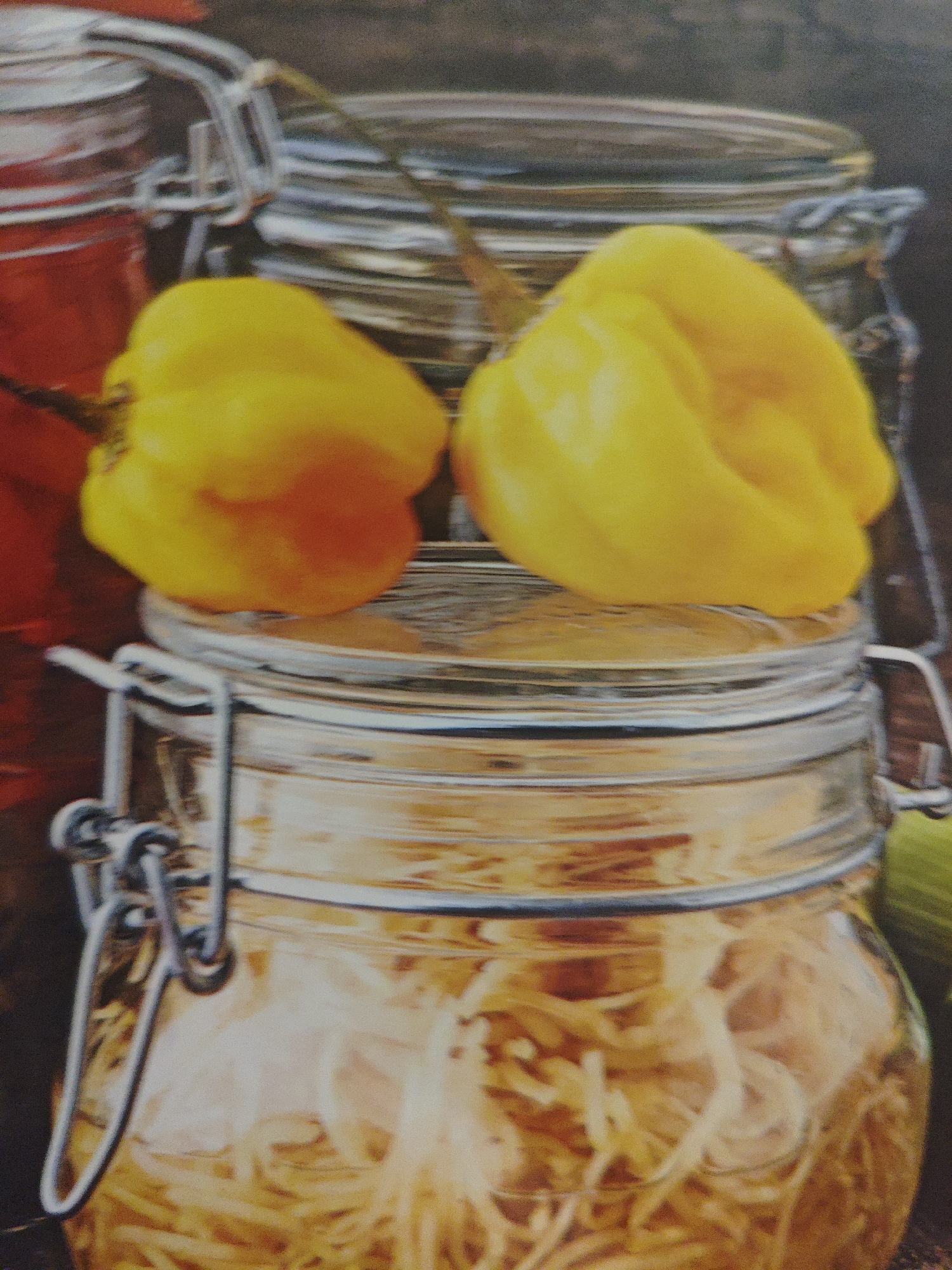 In the foreground of the image, a glass jar with a silver locking mechanism is filled with pale, translucent ingredients, possibly pickled vegetables or fruits. Two bright yellow peppers, likely Habaneros, sit atop this jar. Behind it, another glass jar, also with a silver locking mechanism, is visible, containing a red substance, likely a preserved fruit or vegetable. The background appears to be a dark, textured surface, possibly wood. The image is a close-up shot, focusing on the jars and peppers, with no discernible elements related to the city of Armavir, Russia.