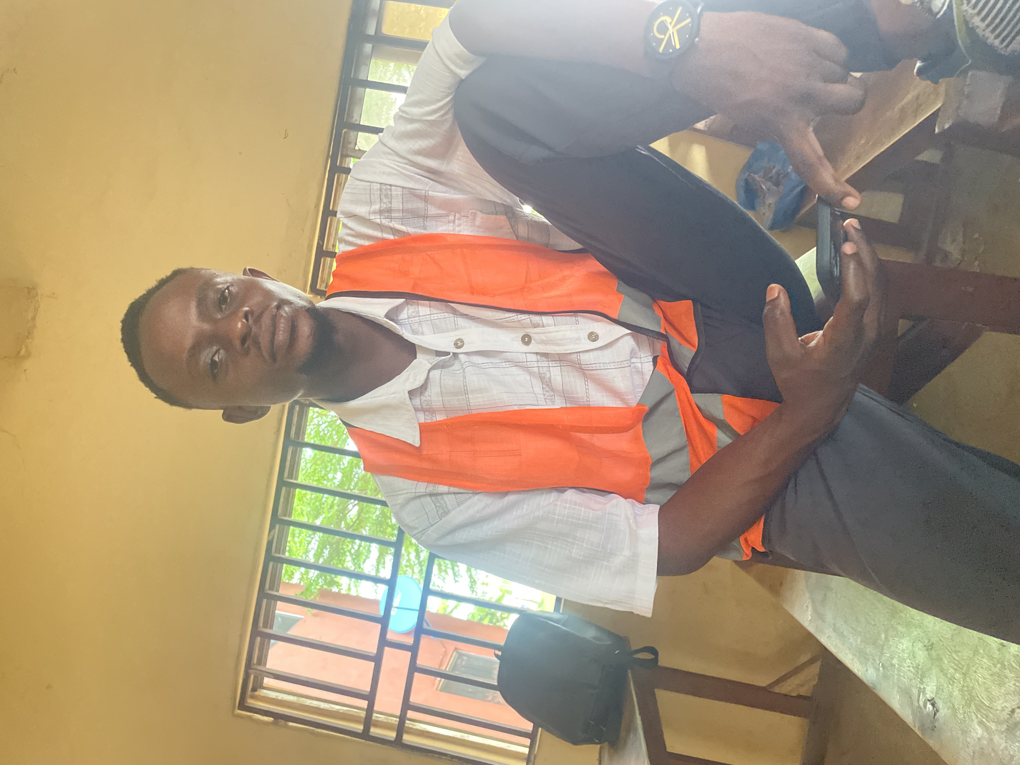 A young Black man wearing a white checkered shirt and an orange safety vest is sitting on a wooden bench in what appears to be a classroom or community center in Lokoja, Nigeria. He is looking towards the camera with a neutral expression. His right leg is crossed over his left, and he is holding a mobile phone in his left hand. His right hand is resting on his right leg. Behind him, a window with metal bars reveals a glimpse of trees and a building. A black backpack is placed on a table to his left. The room has beige walls and the lighting suggests it is daytime.