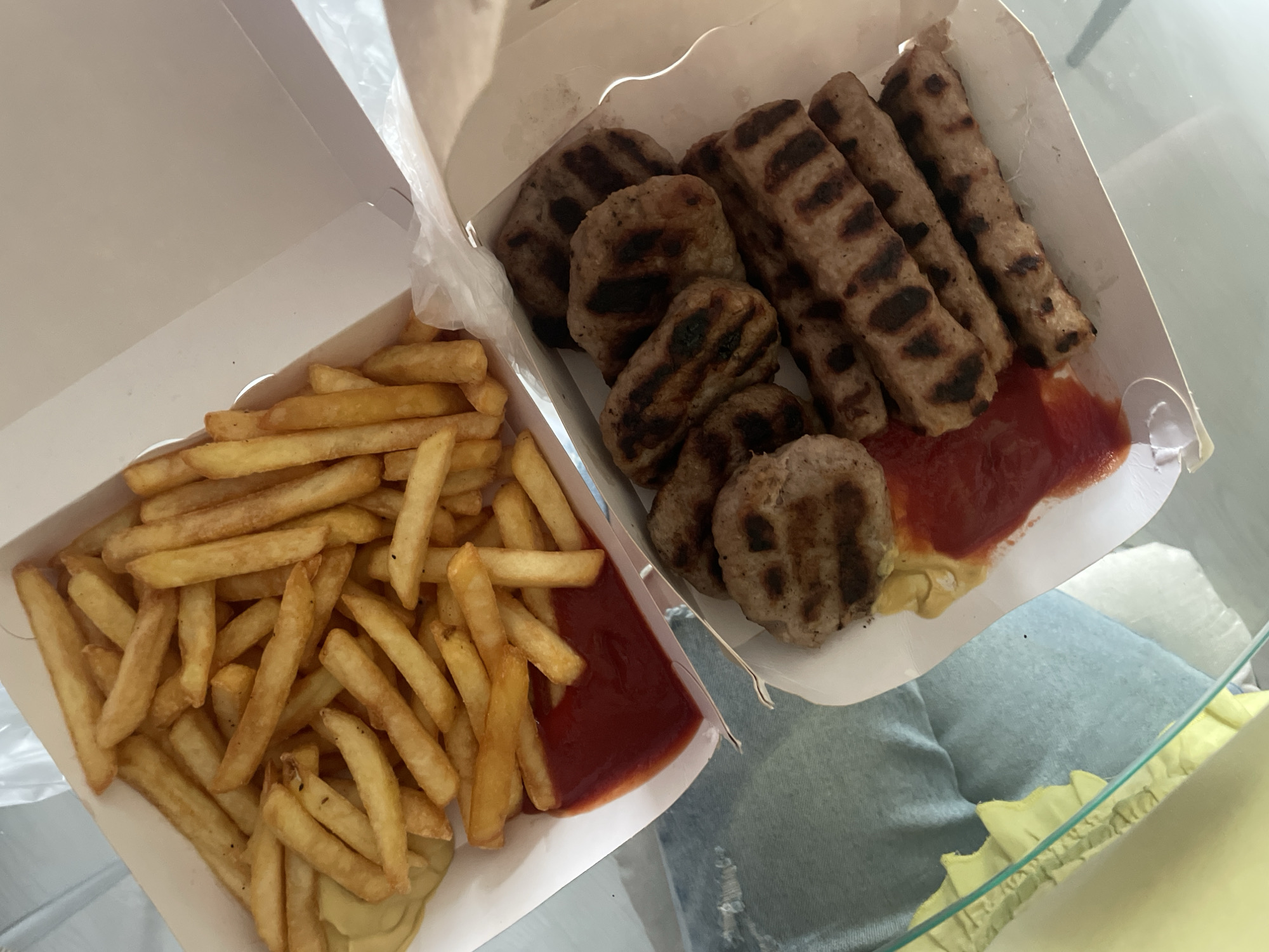 Two white cardboard take-out boxes filled with food are captured from an overhead perspective, resting on a clear, reflective surface, likely a glass table. The setting appears to be indoors.
The box on the left is filled with a large portion of golden-brown french fries. At the bottom right corner of this box, there are generous dollops of red ketchup and a lighter, yellowish sauce, likely mustard.
The box on the right contains various grilled meat items. There are several elongated, cylindrical pieces with distinct char marks, resembling ćevapi. Alongside these, there are flatter, round or oval-shaped patties, also bearing grill marks, which could be pljeskavica or similar grilled meat patties. This box also features a pool of red ketchup and a lighter yellow sauce similar to those in the fries box. A crumpled clear plastic bag is partially visible between and behind the boxes, suggesting the food was recently delivered or picked up.
In the blurred background, partially visible under the glass table, are the legs of a person wearing light blue denim jeans. A hint of yellow, possibly a ruffled fabric, can also be seen below the table line. The types of grilled meat, such as ćevapi and pljeskavica, are common in Balkan cuisine, aligning with the location context of Sofia, Bulgaria. No text is visible in the image.