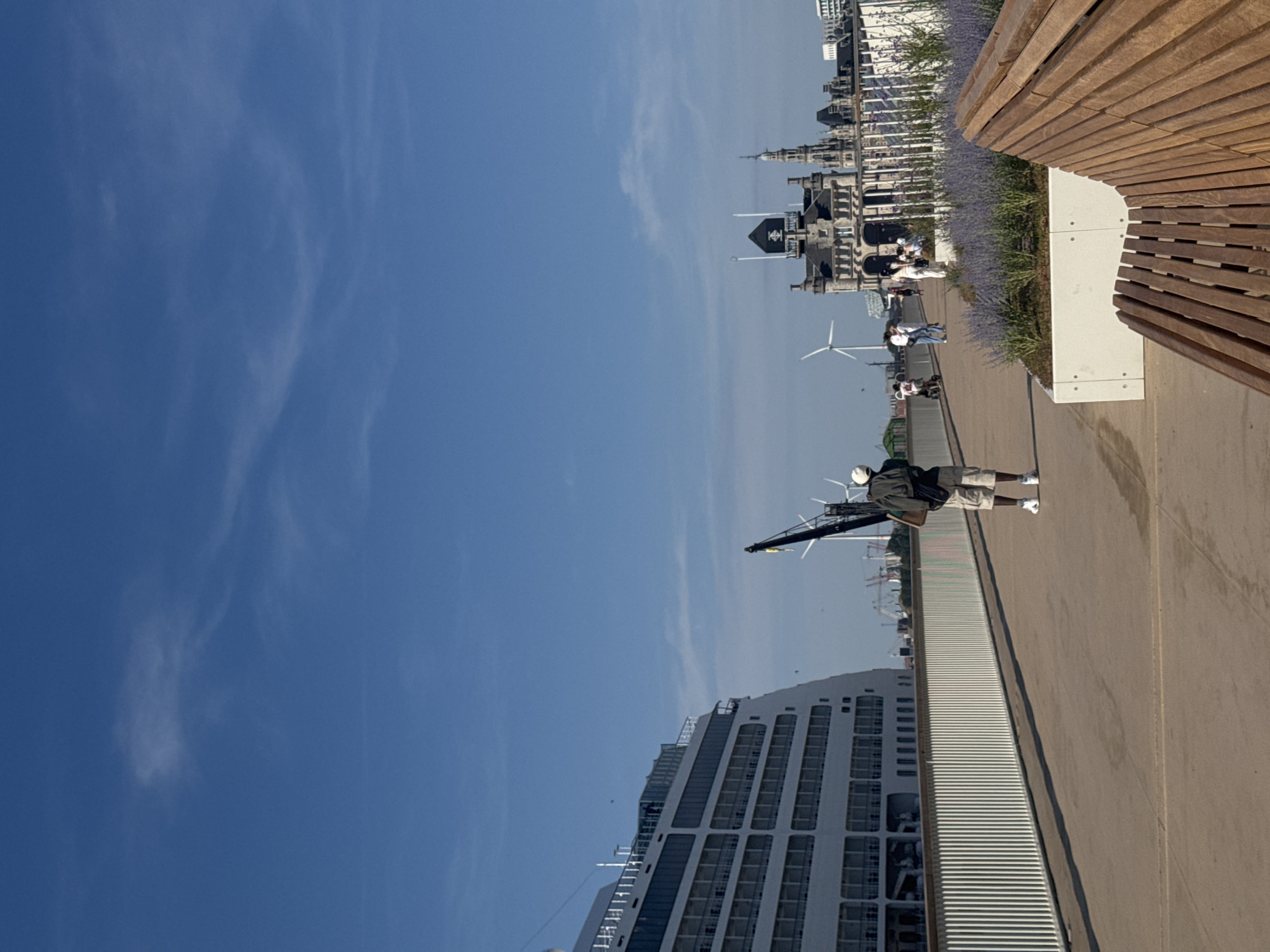 The image depicts a waterfront promenade in the city of Antwerp, Belgium, likely in the port area. A large white cruise ship dominates the left side of the frame, docked alongside a white railing. In the middle ground, a lone individual, a Black man wearing a white cap, a dark jacket, and light-colored shorts, walks away from the camera. Further down the promenade, other people are visible, including a woman on a scooter and a group standing near a building.
The background features industrial elements like cranes and wind turbines, interspersed with ornate historic architecture, notably a building with a distinctive tower and dark facade. Rows of lavender plants are visible in planters along the promenade, adding a touch of color to the scene. A wooden bench is positioned in the foreground, partially obscuring the view of the promenade. The sky is a clear, bright blue with a few wispy clouds. The overall atmosphere suggests a sunny day in a bustling urban port.