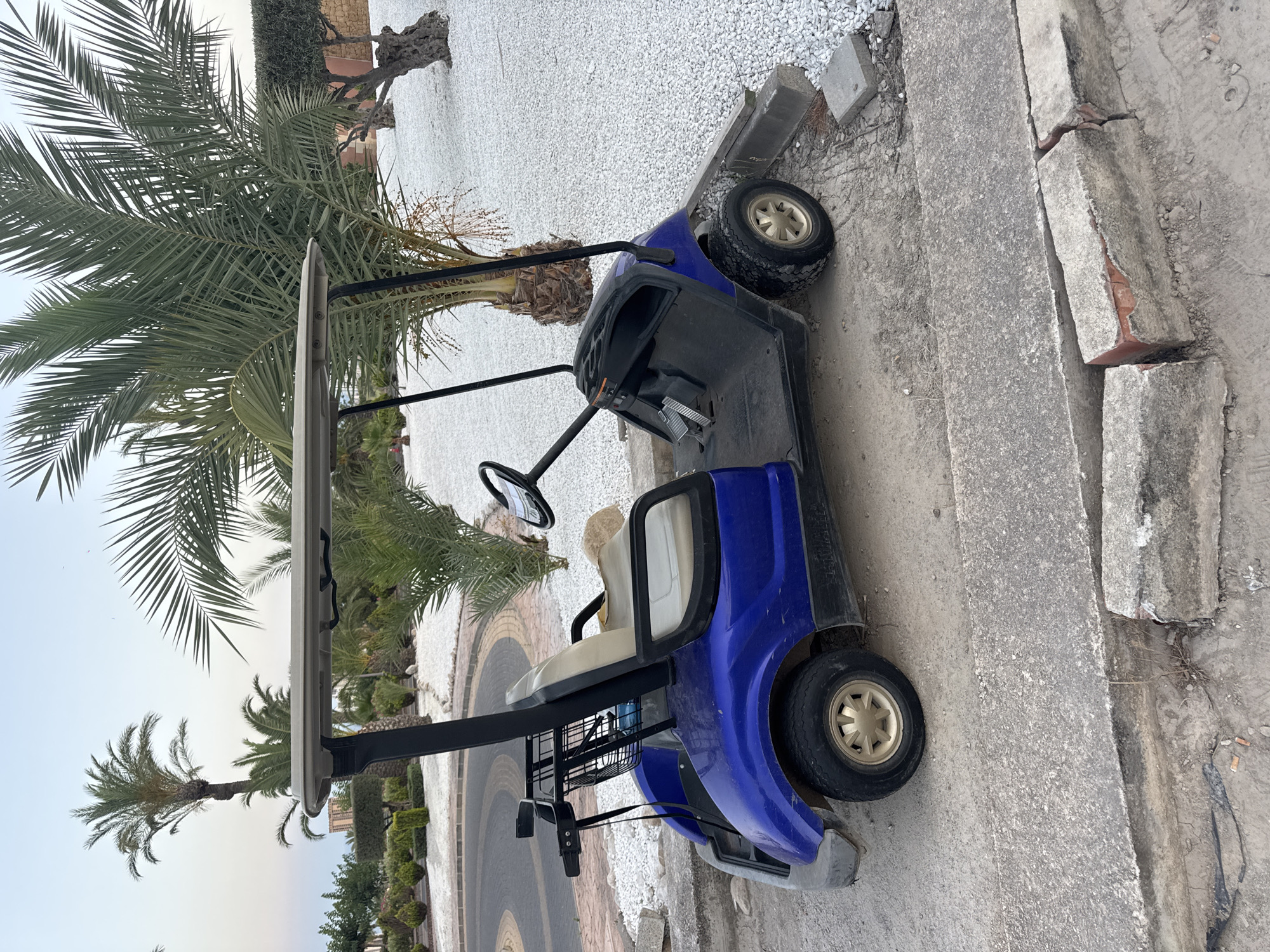 A blue golf cart is parked on a paved surface next to a curb. The golf cart has two seats and a basket on the back. Behind the golf cart, there are palm trees and white gravel. The scene appears to be in a resort or residential area in Benidorm, Spain, given the presence of palm trees and well-maintained landscaping. The sky is clear and a soft blue, suggesting it is either early morning or late evening. The lighting indicates it is not midday.