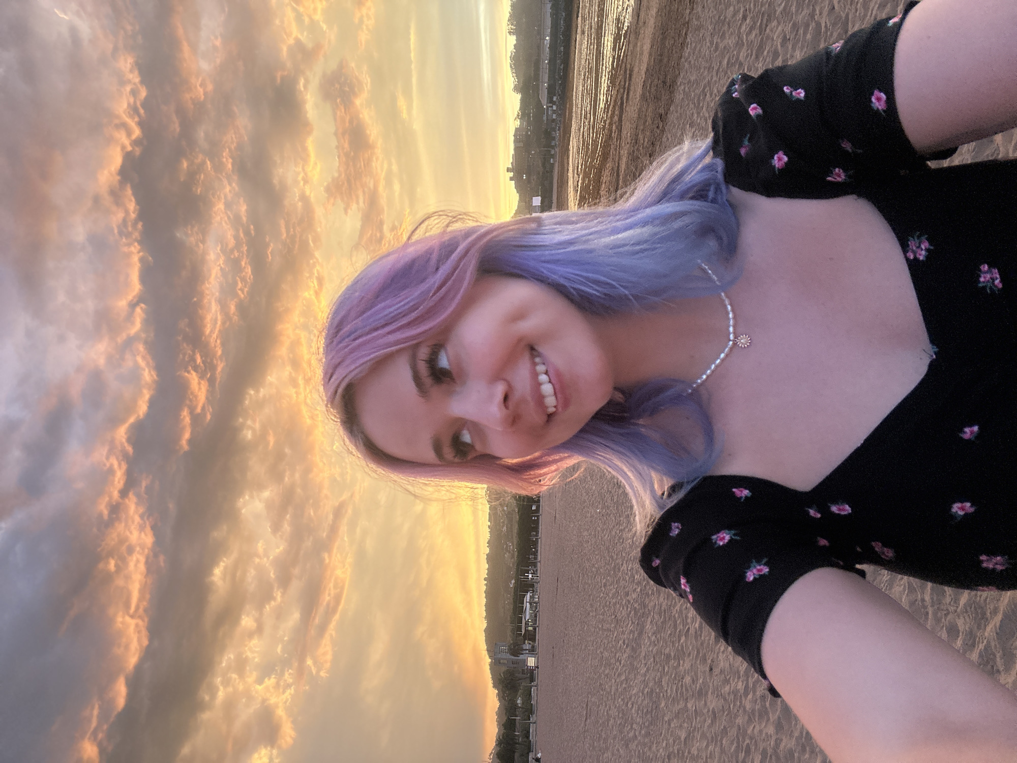A young woman with purple and pink dyed hair takes a selfie on a beach at sunset. She is wearing a black dress with a floral pattern and a pearl necklace. The sky behind her is filled with orange and pink clouds. The beach is sandy and there are some buildings and hills in the background. The scene is likely in the city of Gijón, Spain, given the context of the image. The woman appears to be happy and is looking up and to the side with a smile.