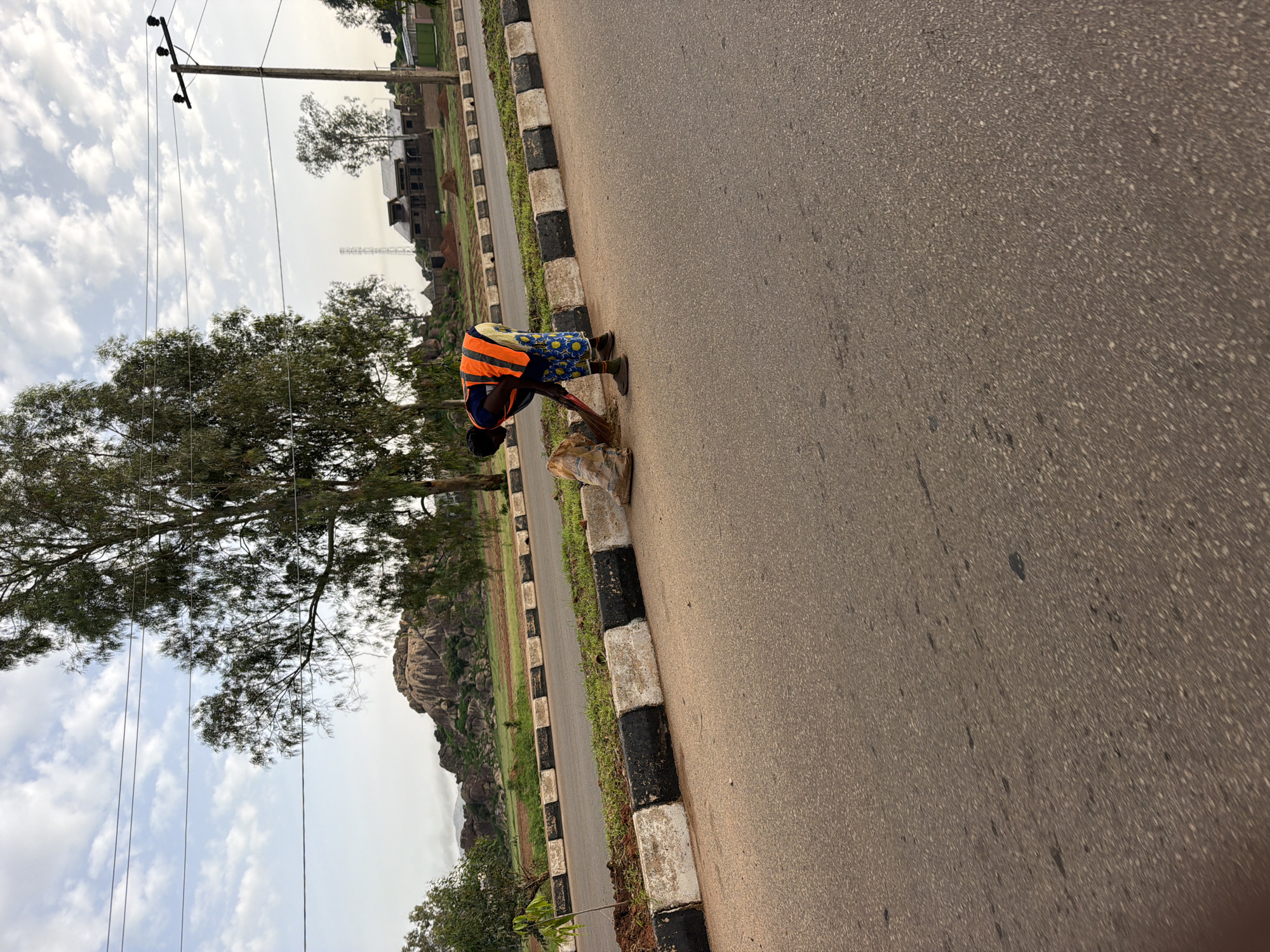 A person wearing an orange high-visibility vest and patterned blue and white pants sweeps debris on the side of a road with a broom. The person is bent over, focused on their task, with a large burlap sack nearby. The road has a concrete curb with alternating black and white sections. In the background, there are trees, a rocky hill, some buildings under construction, and a power line pole. The sky is cloudy. The scene appears to be an outdoor setting in Gura Top, Nigeria.