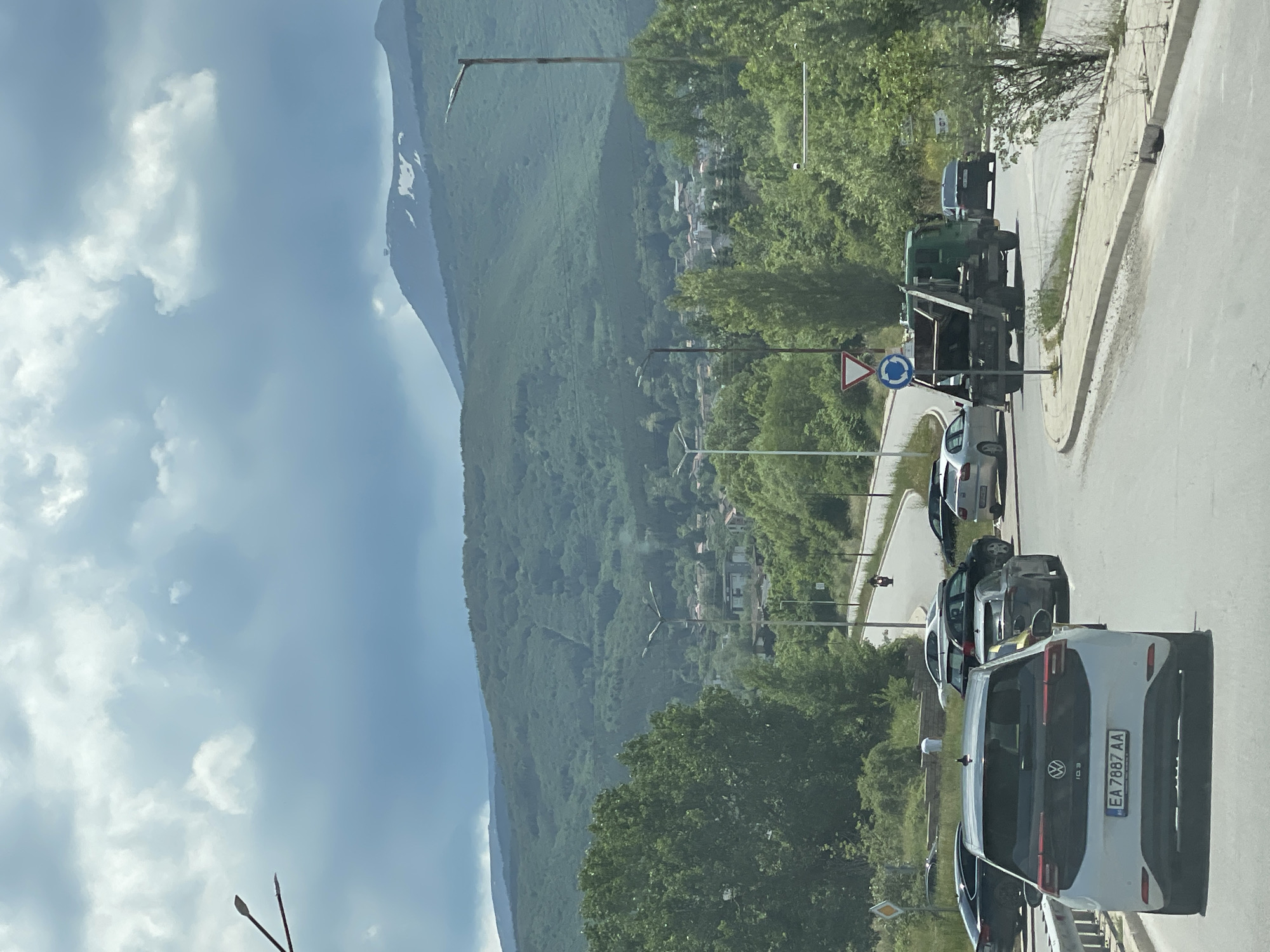 This is a vertical, eye-level, outdoor, daytime, medium shot captured from a vehicle. In the foreground, a white Volkswagen ID.3 electric car with license plate "EA 7887 AA" is prominent, facing away from the camera. To the right, a green garbage truck is turning a corner, with a motorcyclist visible further up the road. Several other cars are also present on the road, some parked and others in motion. The scene unfolds on a winding road, suggesting a suburban or semi-rural setting. Beyond the road, a dense forest covers rolling hills, and in the background, a large, majestic mountain range is visible, with patches of snow on its peak. The sky is partly cloudy, with dramatic gray clouds dominating the upper portion of the frame. The overall impression is a snapshot of traffic and scenery in Sofia, Bulgaria, possibly captured during a scenic drive or commute.