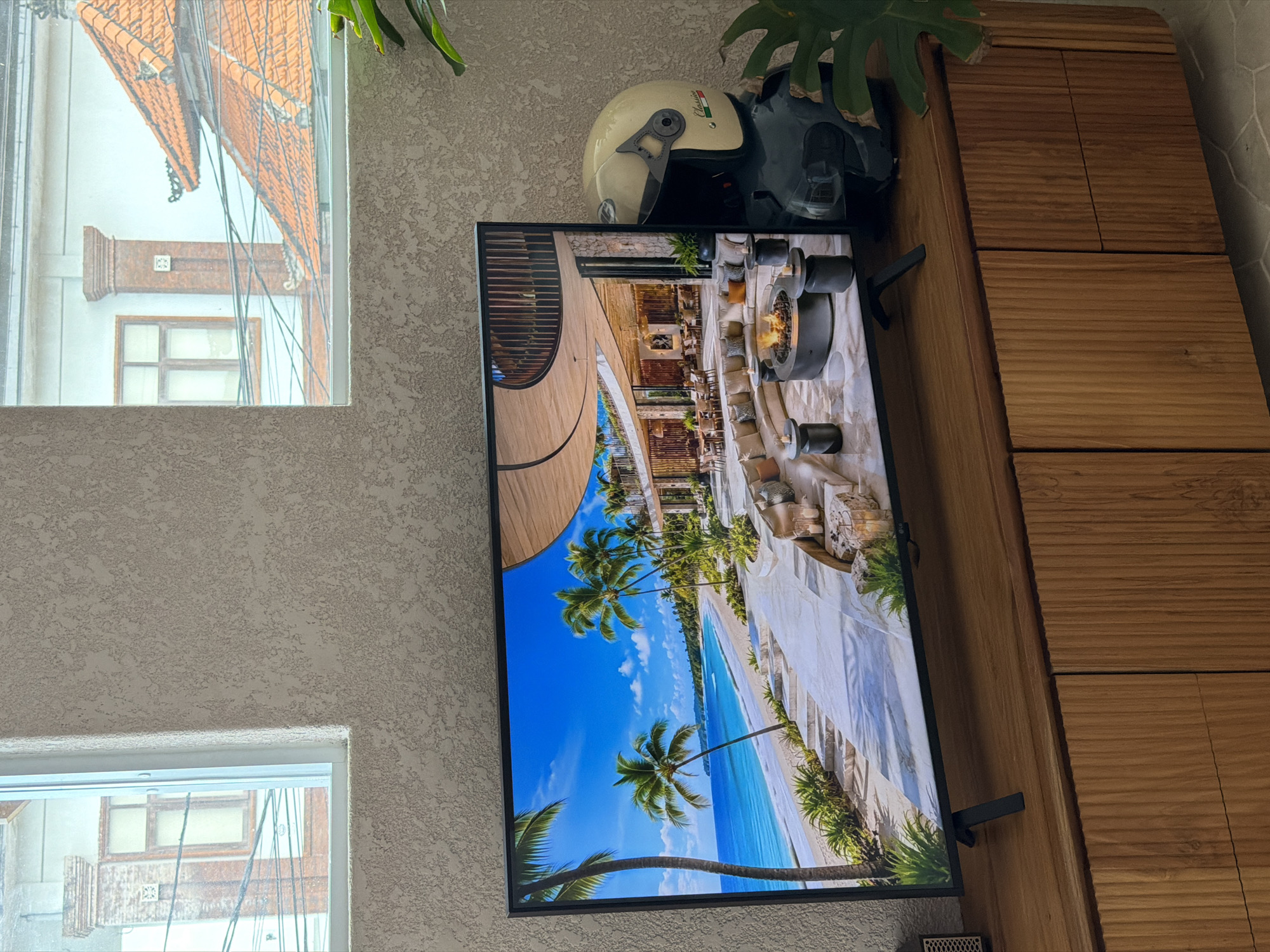 An interior shot shows a large, flat-screen television displaying a tropical beach resort scene. The resort features a white sand beach, turquoise water, palm trees, and modern architecture with an open-air lounge area with a fire pit. To the right of the television, a motorcycle helmet, light beige in color with Italian flag accents, rests on a dark surface. The television and helmet are situated on a wooden media console. The wall behind the television has a textured, light-colored finish. Above the television, two windows offer a glimpse of the exterior, revealing traditional Balinese-style rooftops, brick pillars, and utility wires, suggesting a location in or around Mengwi, Indonesia. A large green plant is partially visible in the bottom right corner.