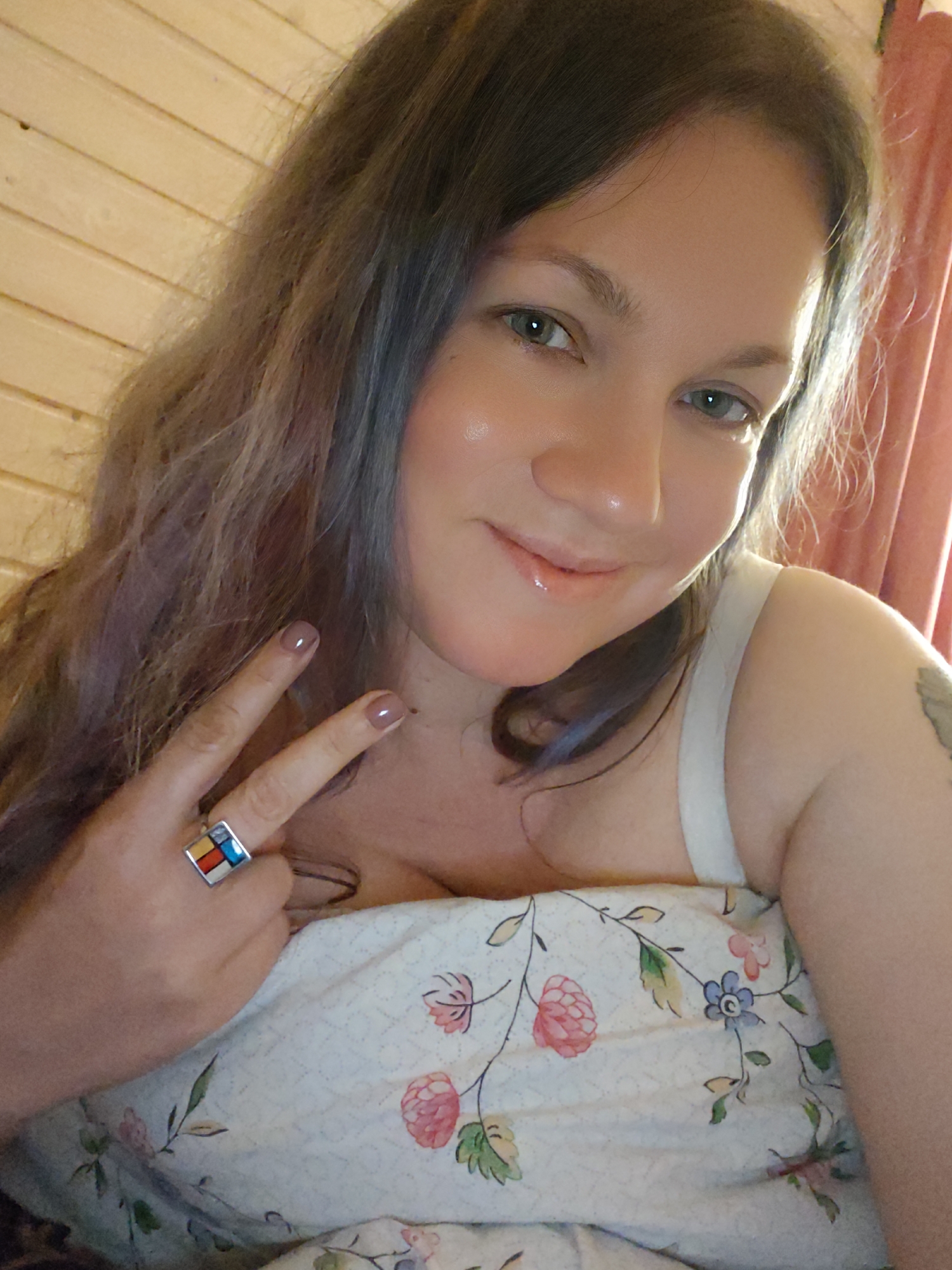 A close-up indoor selfie features a woman with long, dark hair with reddish-purple highlights, smiling softly towards the camera. She has light-colored eyes, glowing skin, and shiny lips. She appears relaxed and is making a peace sign with her left hand, which has light-colored nail polish. A prominent square ring with colorful inlays (red, yellow, blue, and white/silver segments) is visible on her left ring finger.
She is partially covered by a floral-patterned duvet or bedsheet featuring pink, green, and blue flowers and leaves on a light background. A light-colored strap, possibly from a camisole or tank top, is visible on her shoulder, and a tattoo is partially visible on her upper right arm.
The background shows warm-toned wooden paneling on the left, suggesting a rustic interior, possibly a cabin or a room with wooden walls. A warm, reddish-orange curtain is visible on the right. The lighting suggests natural daylight, possibly from a window nearby. The overall impression is one of contentment. No text is visible in the image. The scene is set in what is identified as Свистуха (кузяевский С/о), Russia.