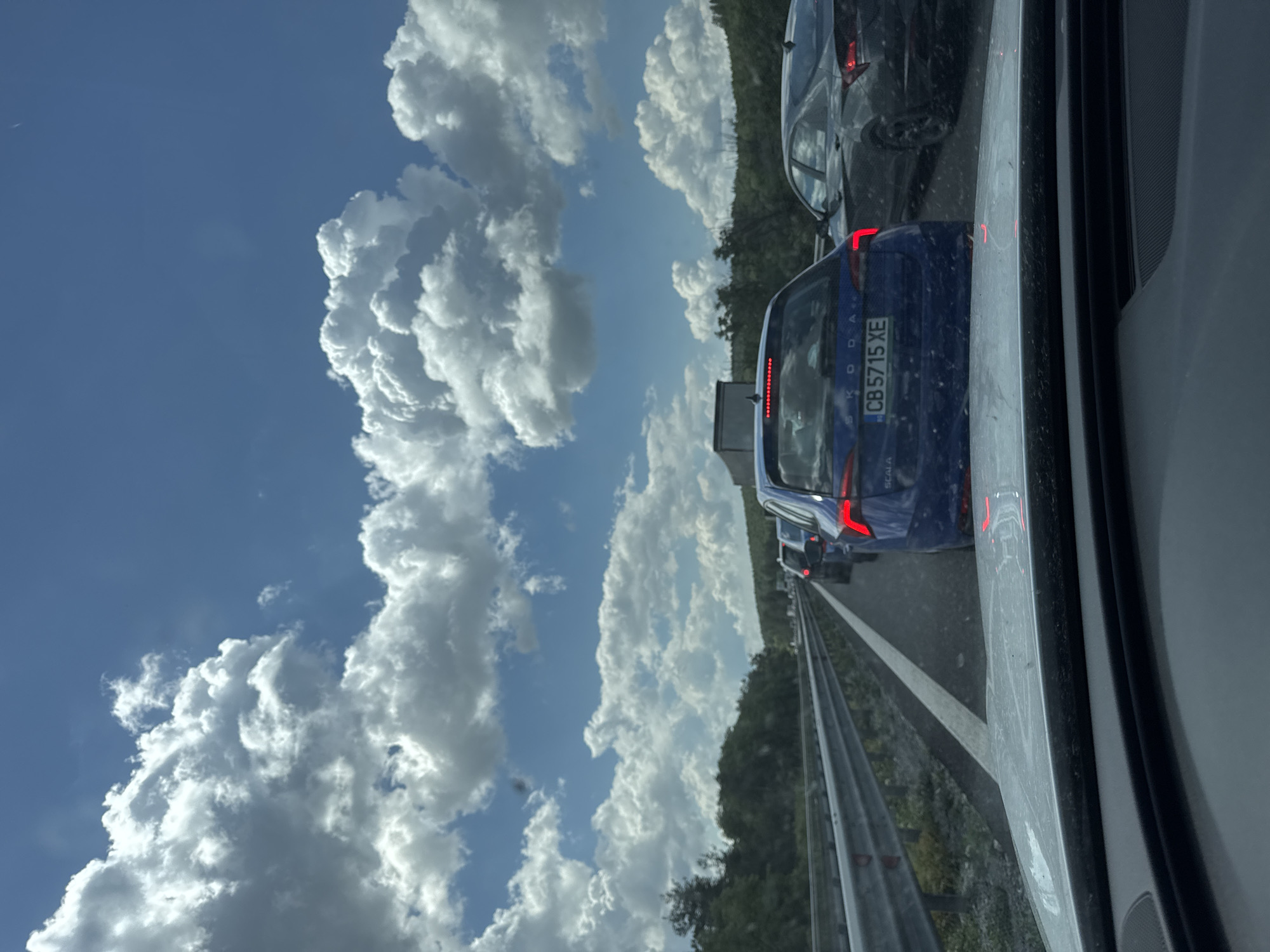 The image shows a traffic jam on a highway. The viewpoint is from inside a vehicle, looking forward. In the foreground, the dashboard and windshield of the vehicle are visible. Beyond the windshield, several cars are stopped in traffic, including a blue Skoda with license plate CB 5715 XE. The road is asphalt, with a metal guardrail on the left side. On either side of the highway, there are trees and vegetation. The sky is blue with white, fluffy clouds. The scene appears to be in Rajana, Bulgaria, given the license plate format.