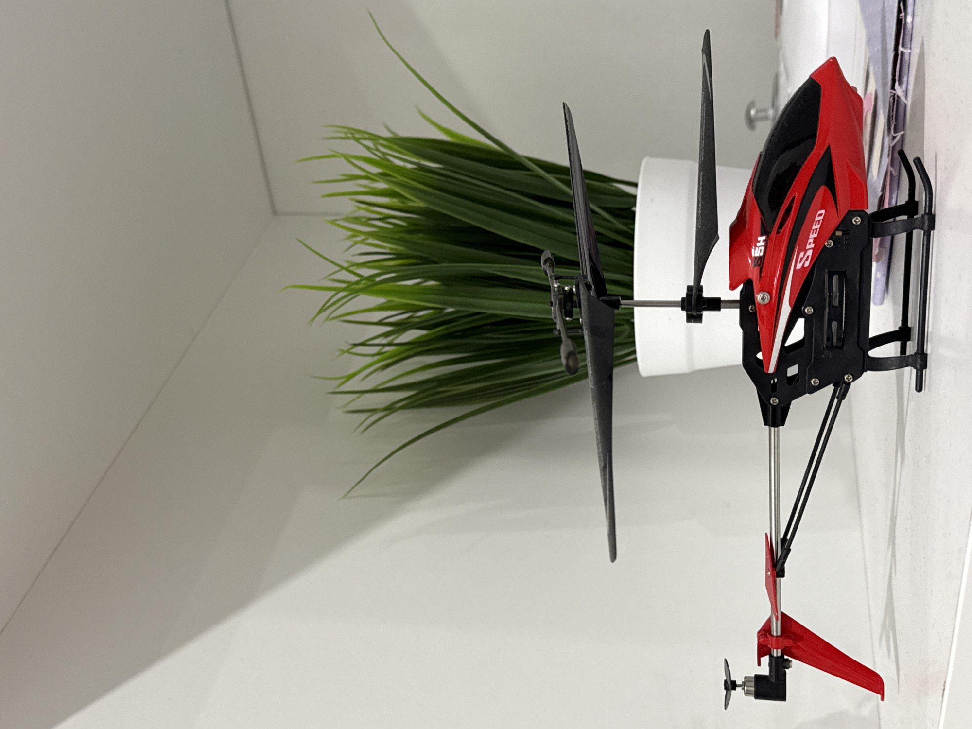 A red and black toy helicopter rests on a white surface, likely a shelf or desk, in an indoor setting. The helicopter's body is predominantly red with black details and white stripes, prominently displaying the word "SPEED" and "5H" on its side. It features two black main rotor blades and a smaller red tail fin with a black tail rotor assembly.
Behind the helicopter, a white potted plant with artificial green, grass-like foliage adds a touch of color to the scene. The plant is housed in a simple white cylindrical pot. The background consists of plain white walls and surfaces, suggesting a clean and minimalist environment. The lighting is bright and even, casting soft shadows, indicative of diffused indoor illumination. No activity is taking place, and no people are visible. The scene is static. While the location context of Yekaterinburg, Russia, is provided, there are no specific visual cues in the image to confirm this.