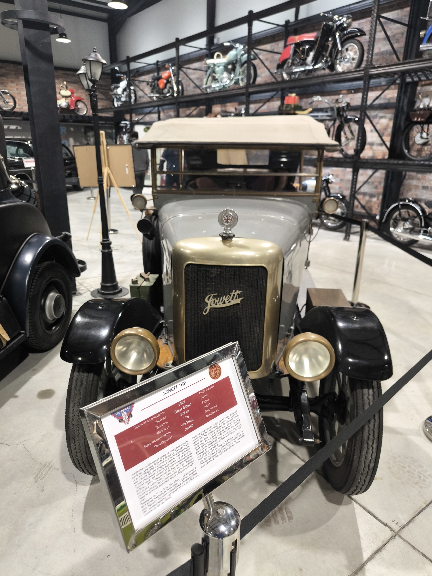 The image shows a grey 1927 Jowett 7HP car on display. The car has black fenders and a gold-colored grill with the word "Jowett" on it. A display stand with information about the car is placed in front. The background reveals a museum-like setting with vintage motorcycles displayed on racks. The location is implied to be Kapatovo, Bulgaria, based on the text on the display stand.