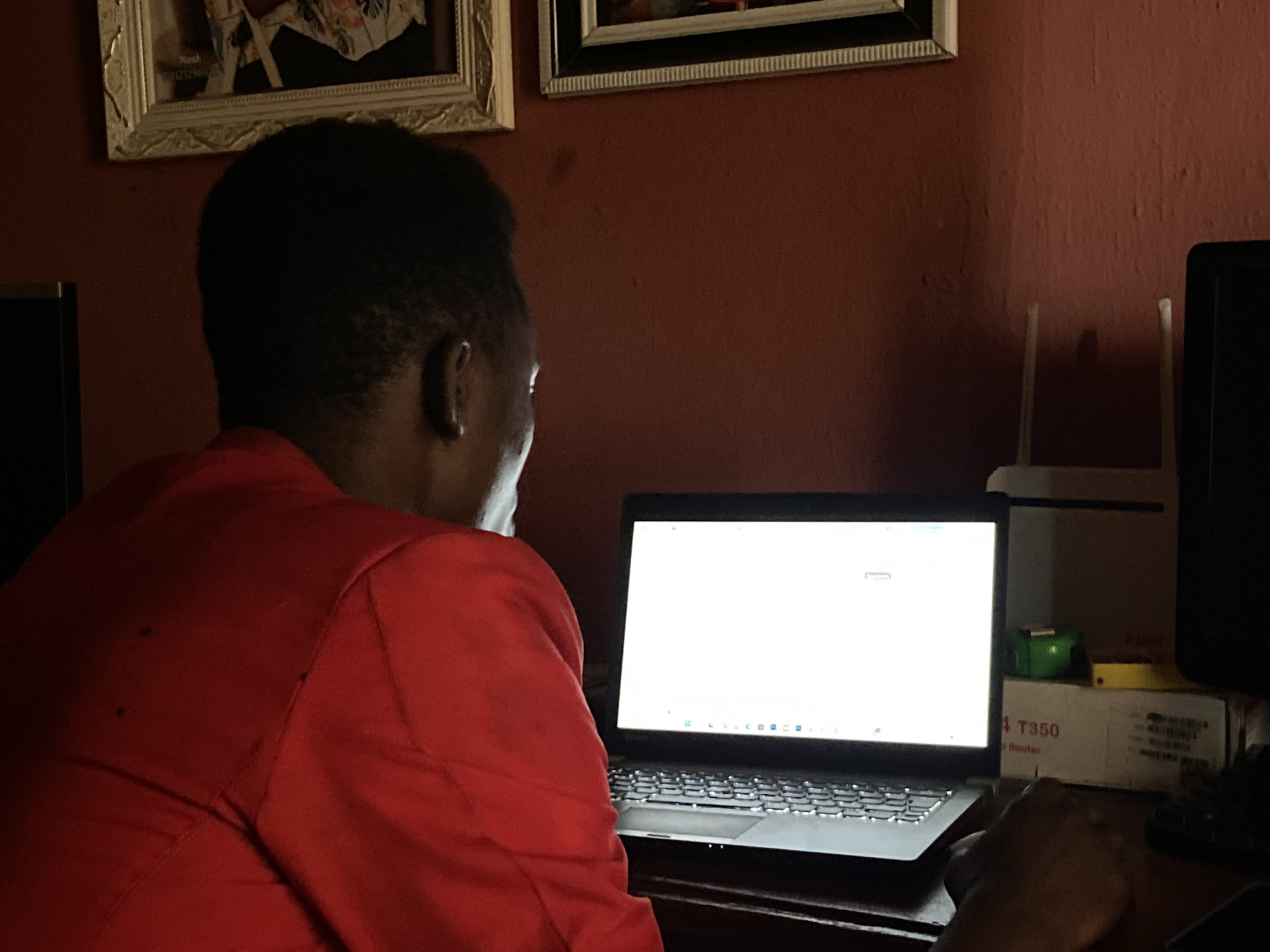 An individual is depicted from the back, seated at a desk and interacting with a laptop in an indoor environment in Mararaba, Nigeria. The person, with short dark hair and wearing a red long-sleeved garment, faces a laptop with a brightly illuminated white screen displaying a taskbar with icons at the bottom. The individual's right hand rests on a dark computer mouse positioned on a dark wooden desk surface. To the right of the laptop, a white router with two antennas is visible, situated on a white cardboard box that has "T350" and "Router" printed on it. A small green object and a thin yellow object are placed on top of this box. The walls of the room are a dark red hue, and two framed pictures, one with an ornate gold frame and another with a black and white striped frame, are mounted on the wall above the individual's head. The laptop screen appears to be the primary light source in the otherwise dimly lit room.
